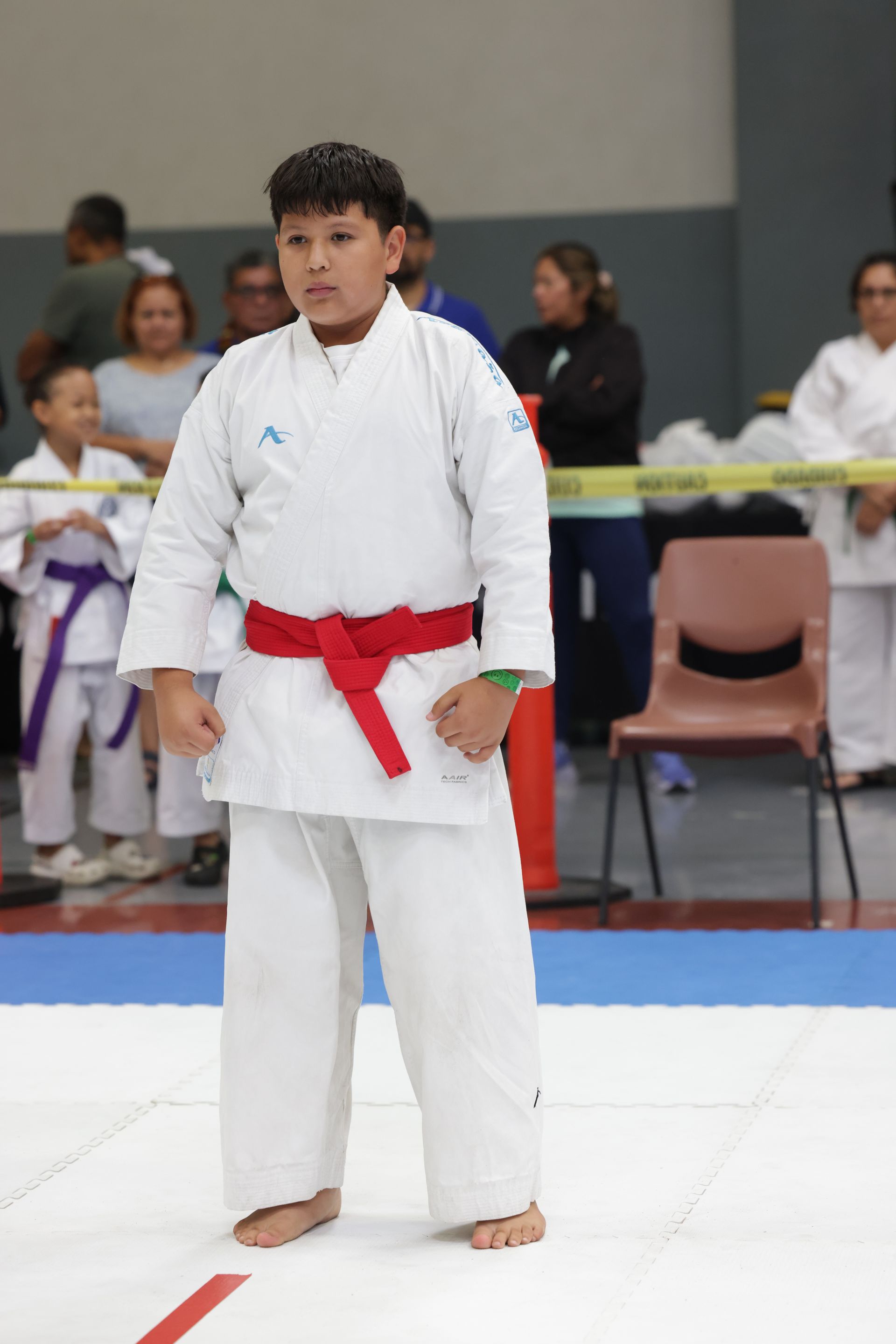 Martial artist in a white gi with red belt stands on a mat at an indoor competition