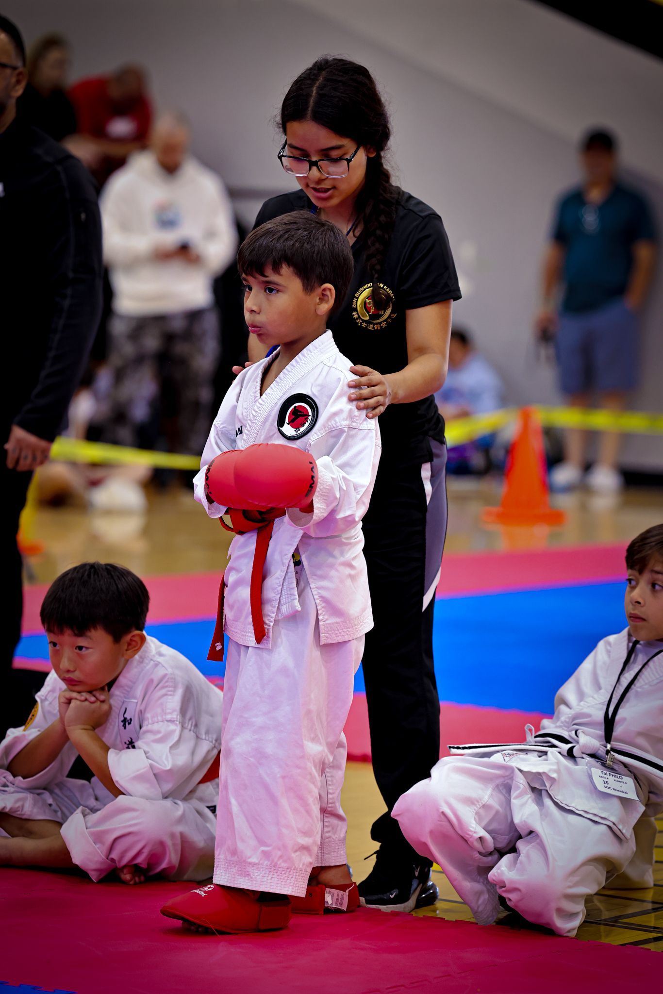 A child in a white karate uniform and red gloves stands with a coach in a gym, with other students sitting nearby.
