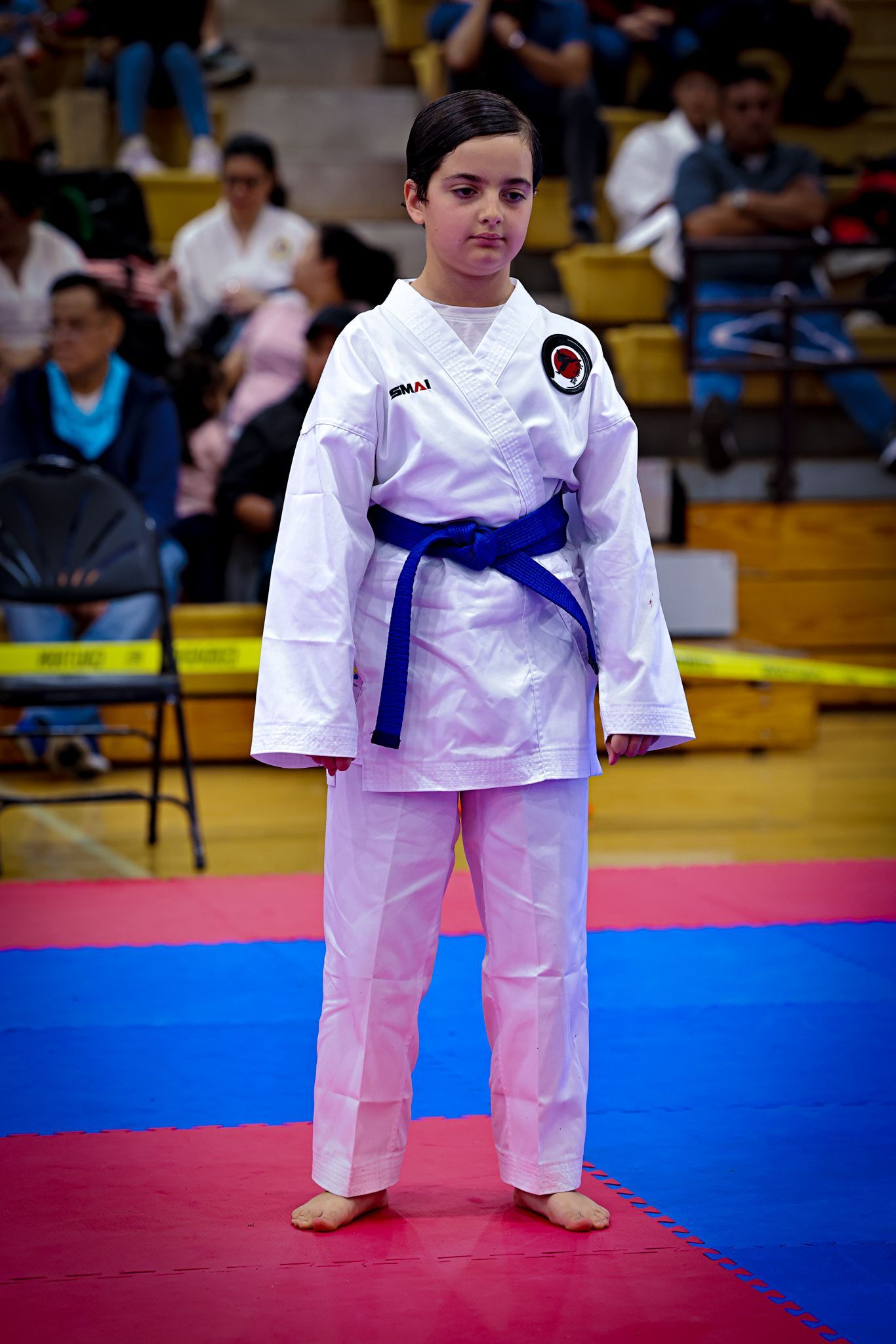 A student in a white karate uniform with a blue belt stands centered on a blue and red mat in a gymnasium.