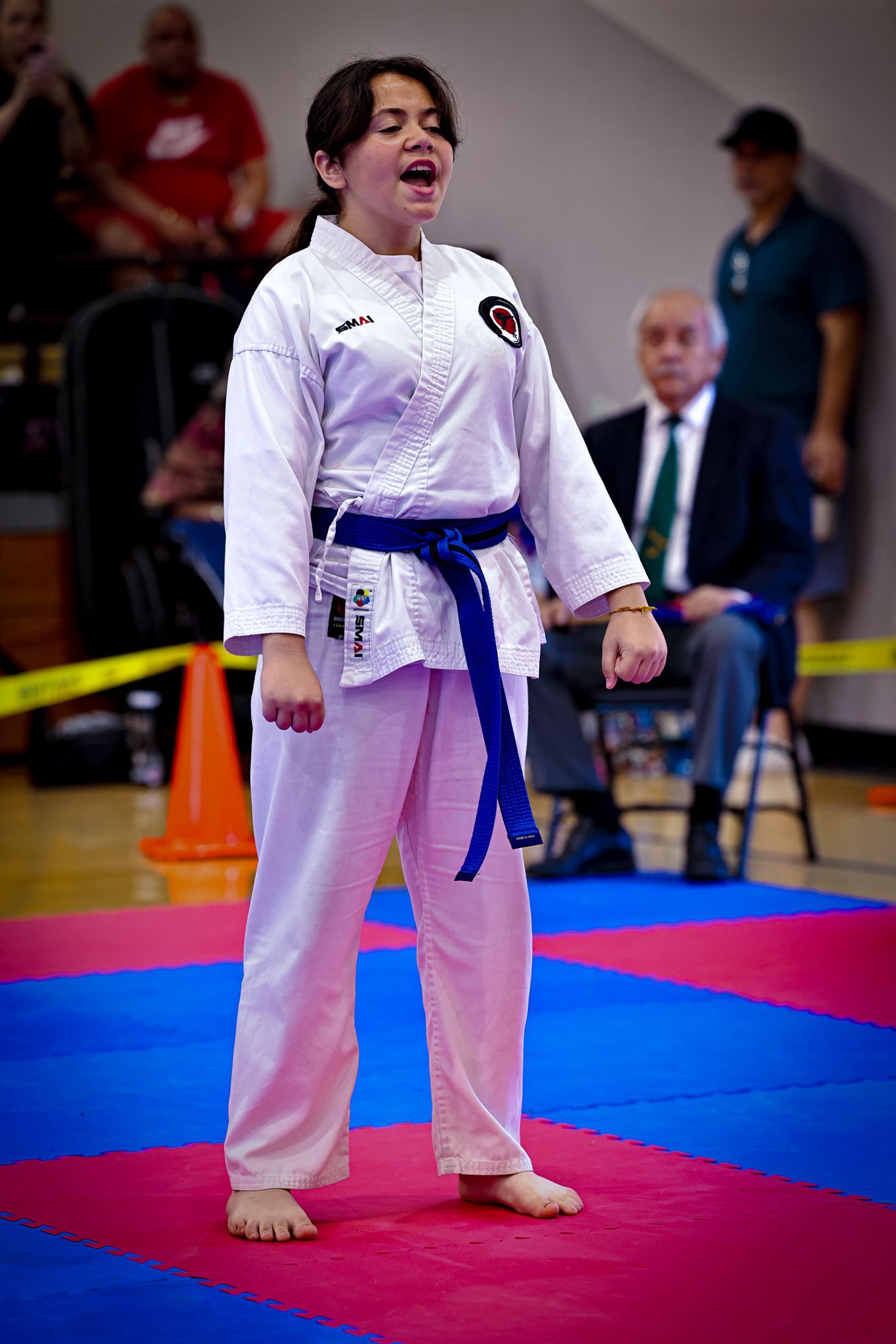 A martial artist in a white uniform with a blue belt stands on a colorful mat, shouting with a focused expression.
