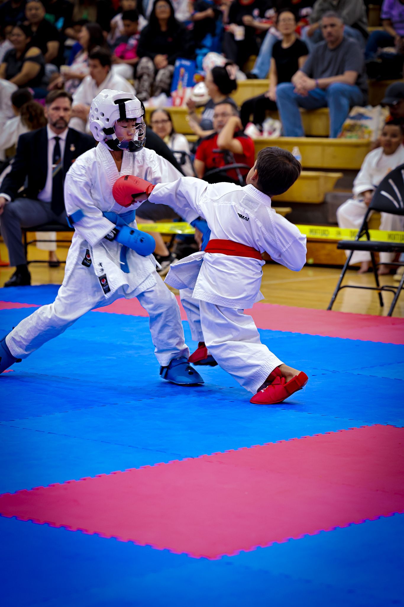 Two karate competitors in white gis sparring on a blue and pink mat in front of a seated audience.