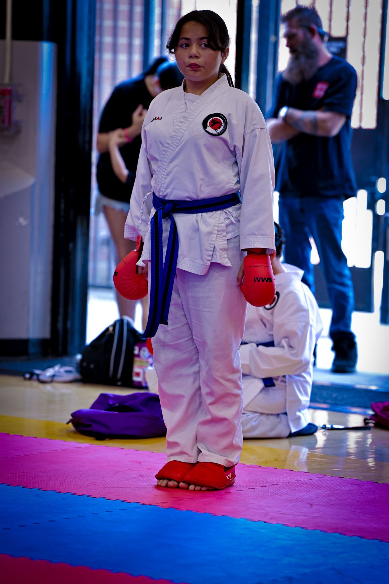 A martial artist stands in a white uniform with a blue belt and red gloves on a multi-colored mat in an indoor gym.
