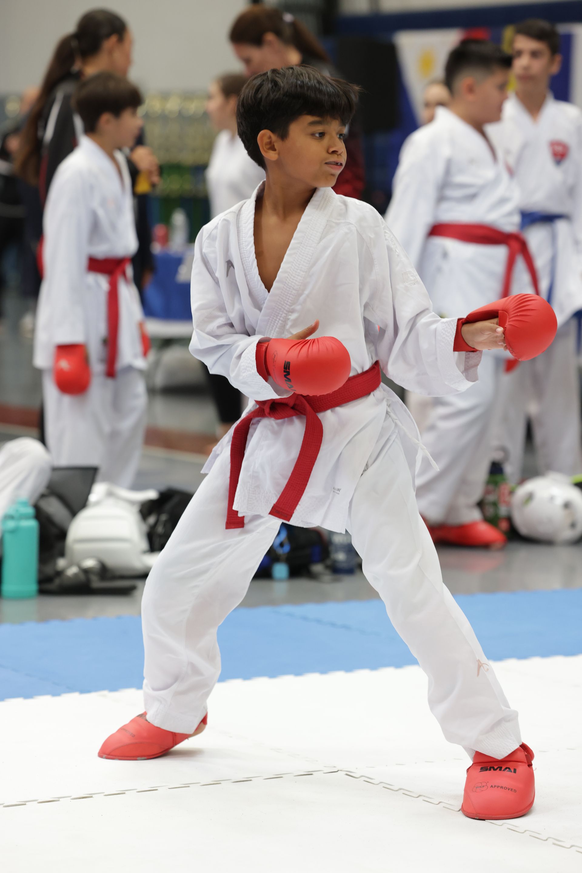 Karate competitor in white gi with red gloves and belt stands in fighting stance on a mat, others blurred behind.