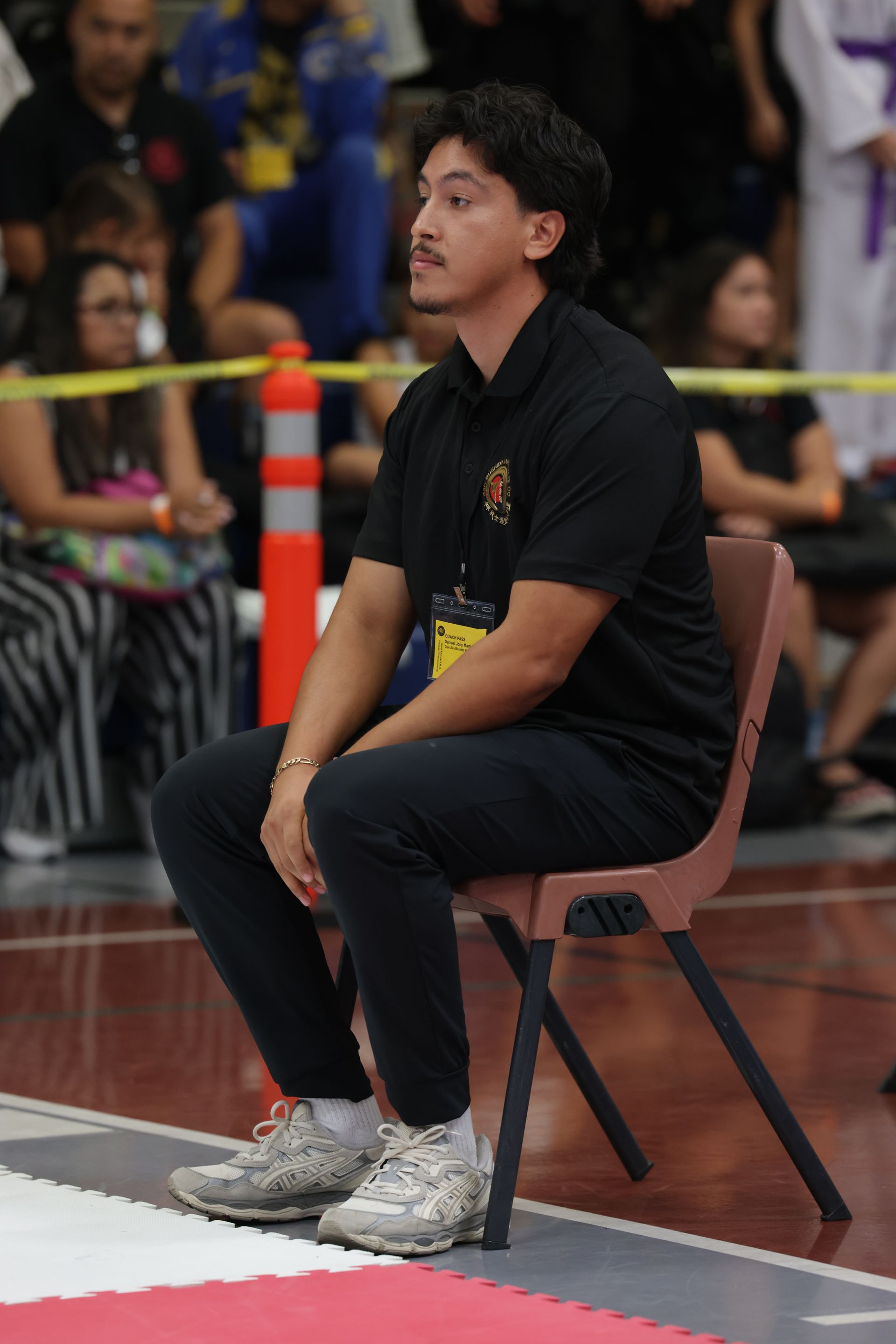 Man sitting on a chair courtside in a black shirt and pants, watching an indoor event.