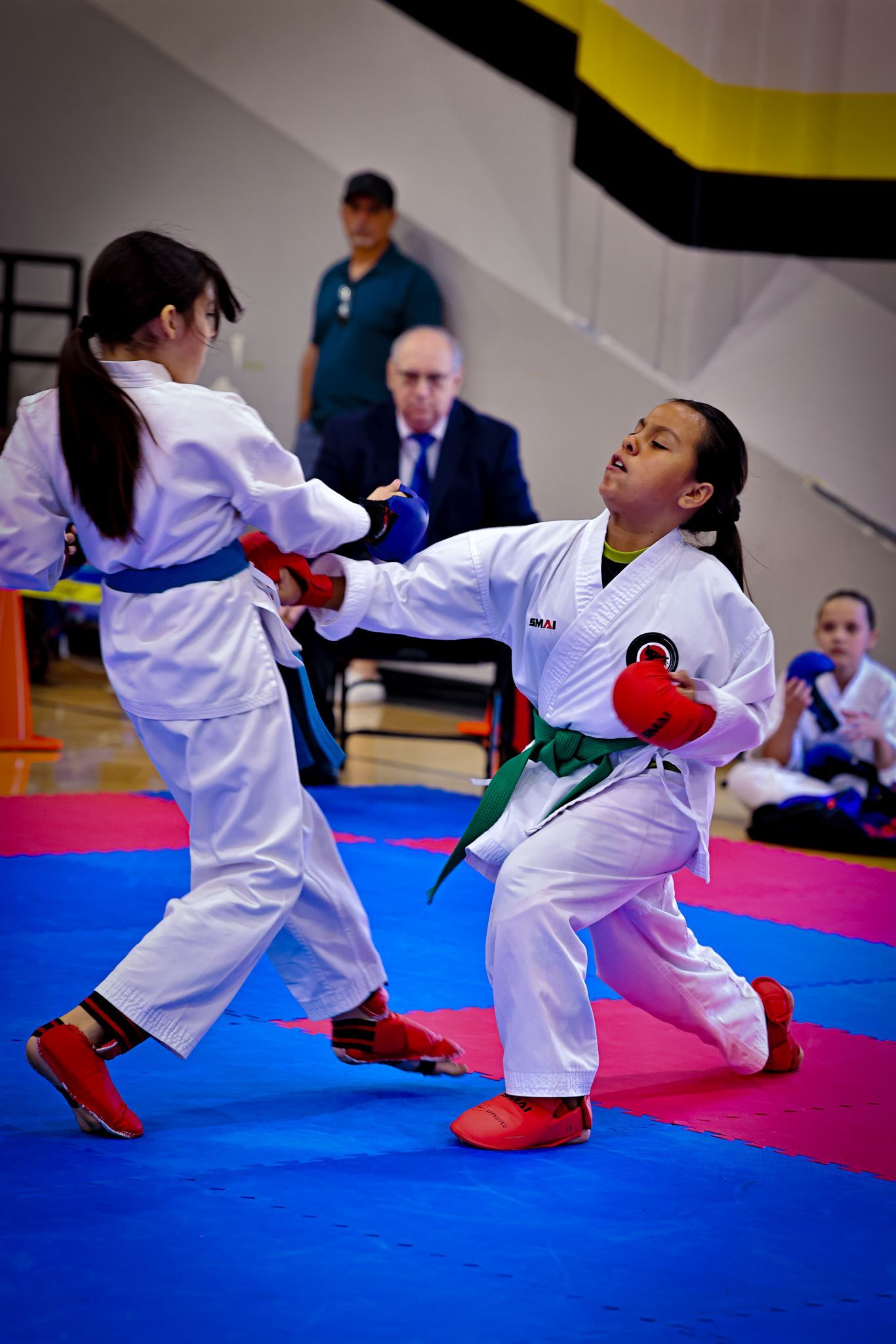 Two karate practitioners in white uniforms with blue and green belts sparring on a mat with red protective gear.