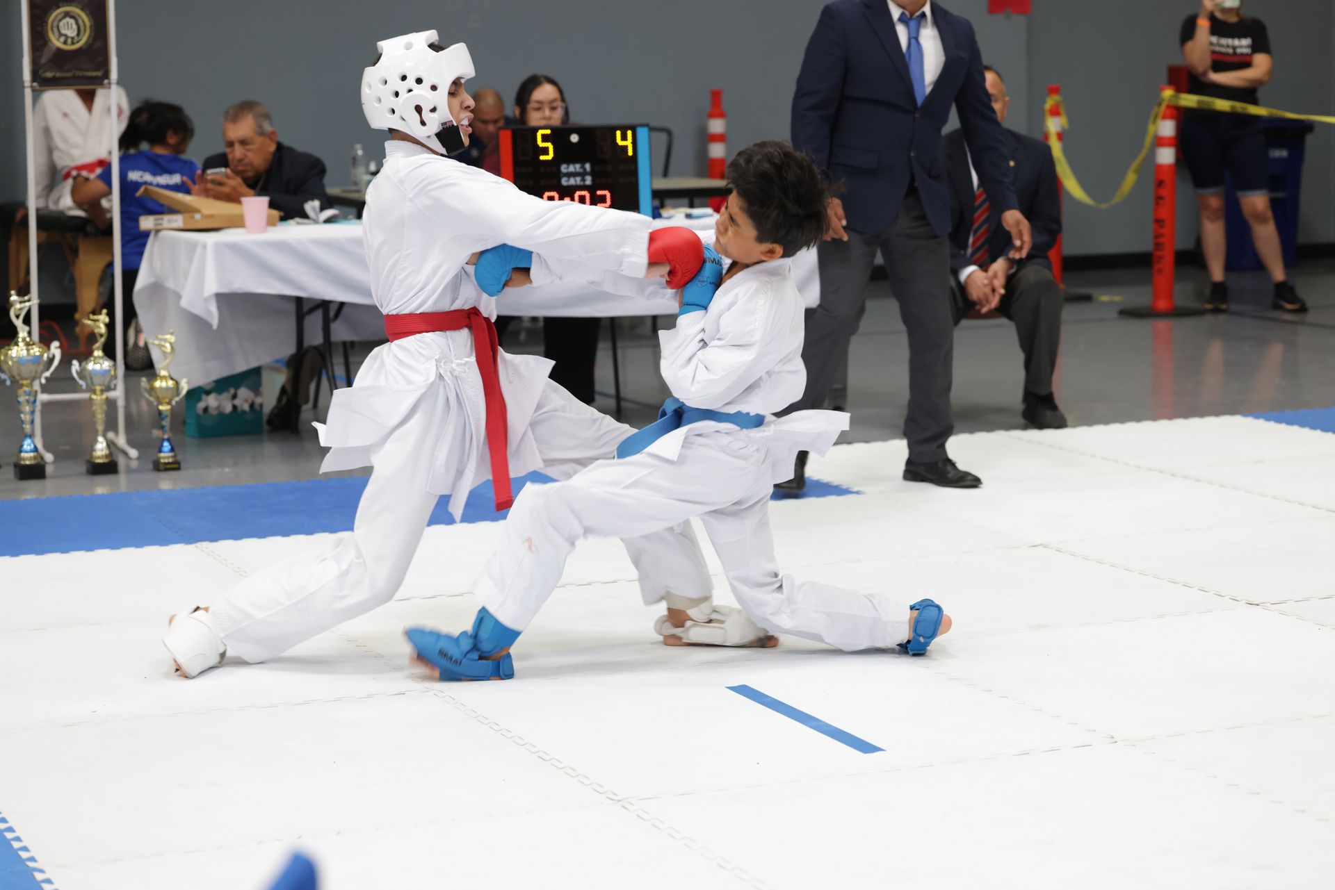 Two martial artists sparring in white uniforms at an indoor competition, one lunging forward with a blue glove.