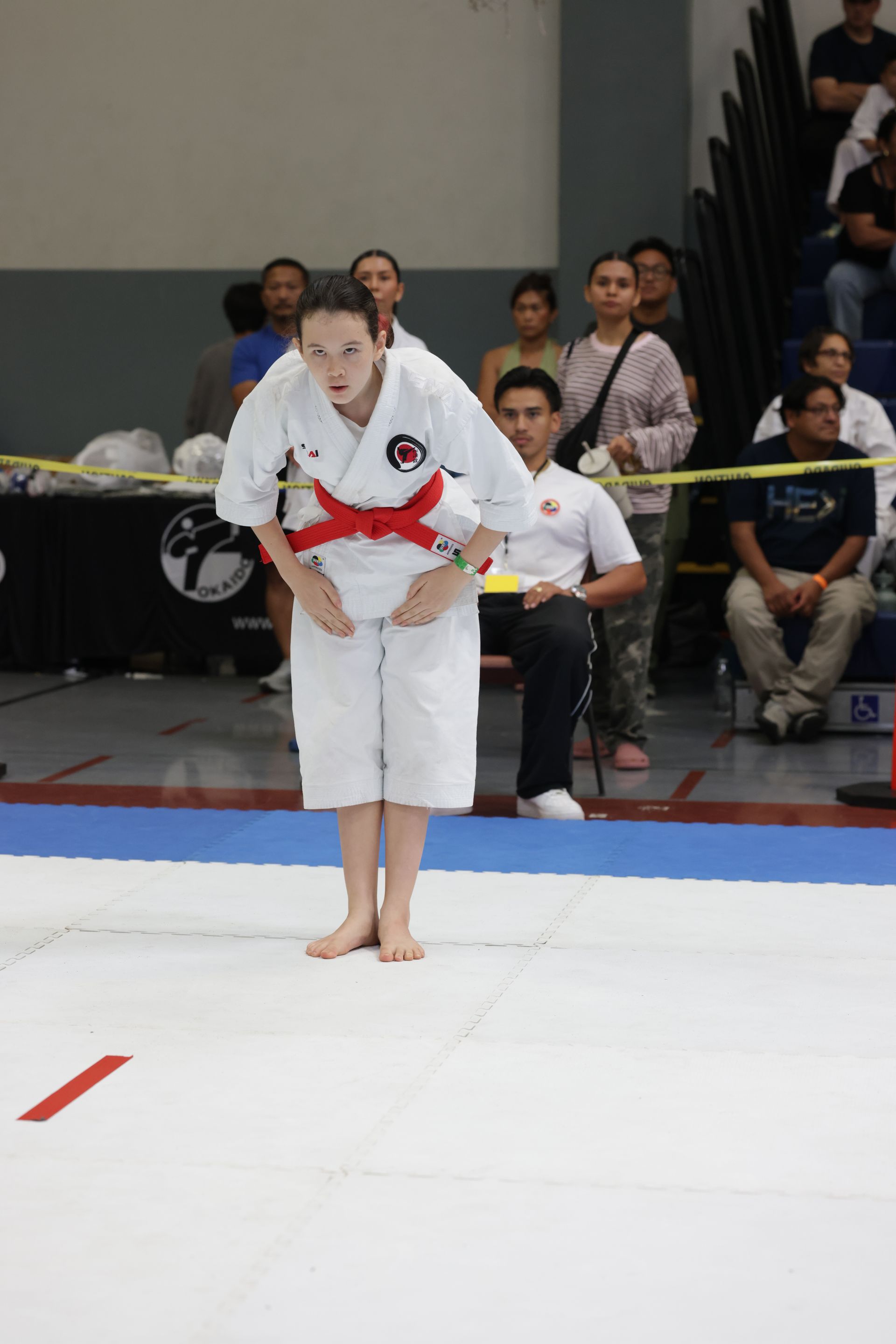 Martial artist in white gi with red belt bows on a mat during a competition, spectators seated behind.