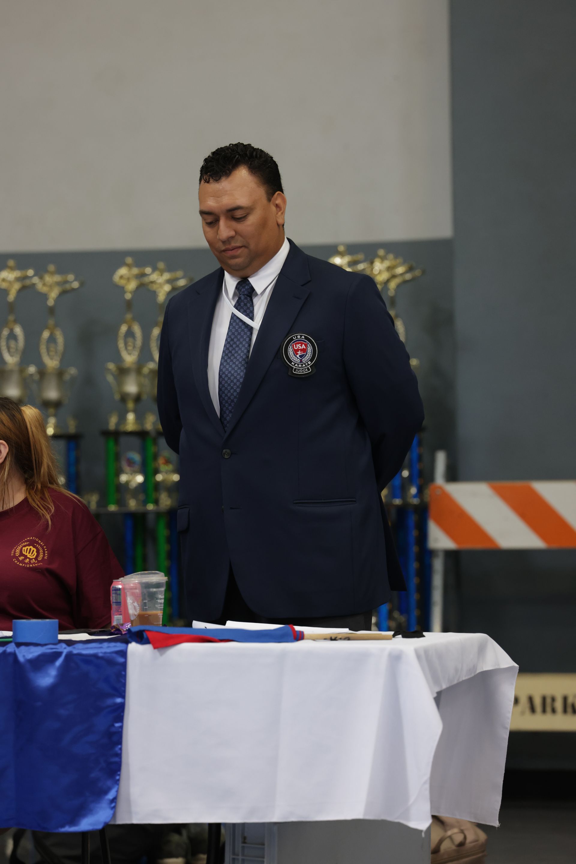 Man in a navy suit stands beside a table with trophies and papers at an indoor event