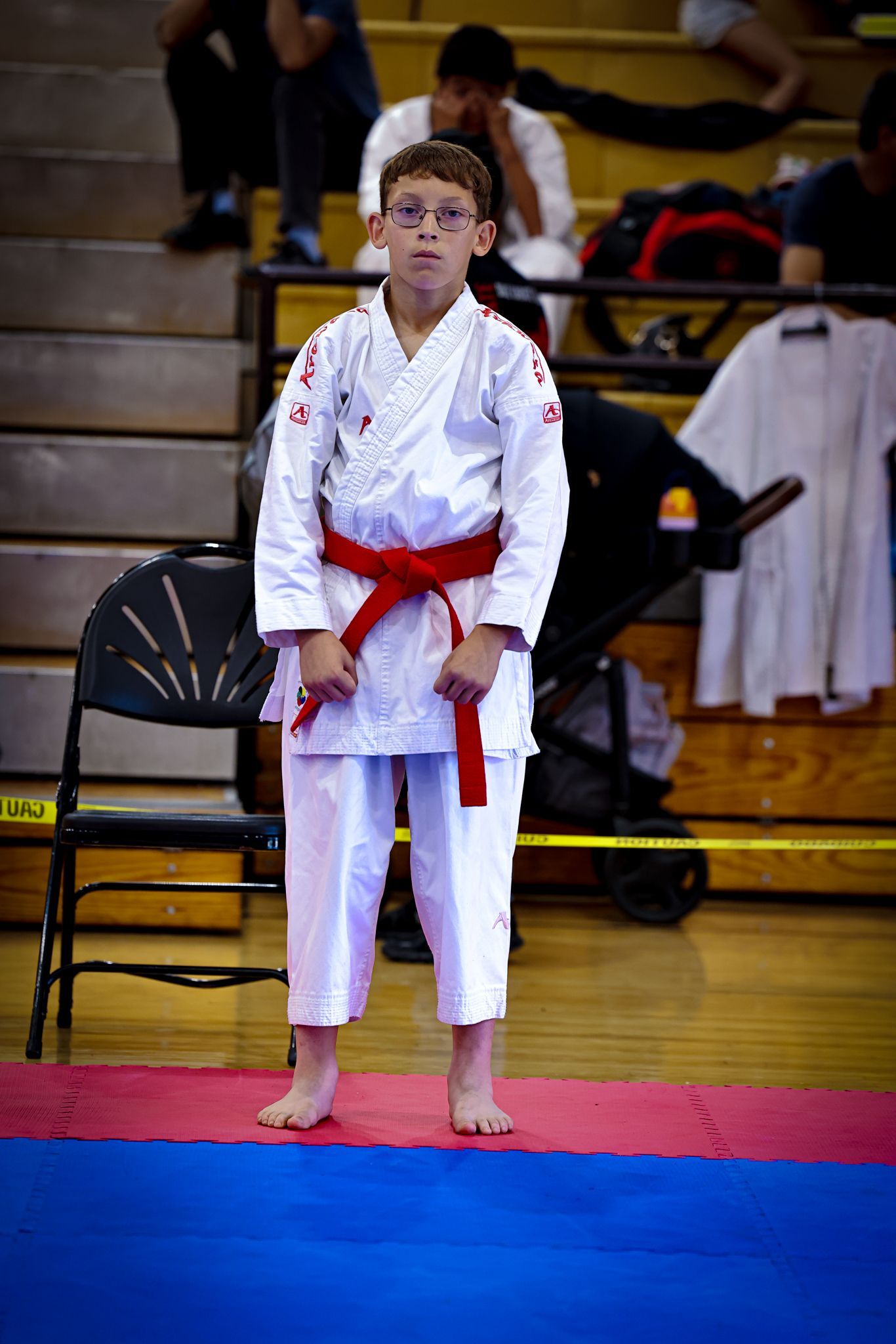 A martial artist with a red belt stands in a white uniform on a blue and red mat in a gymnasium.