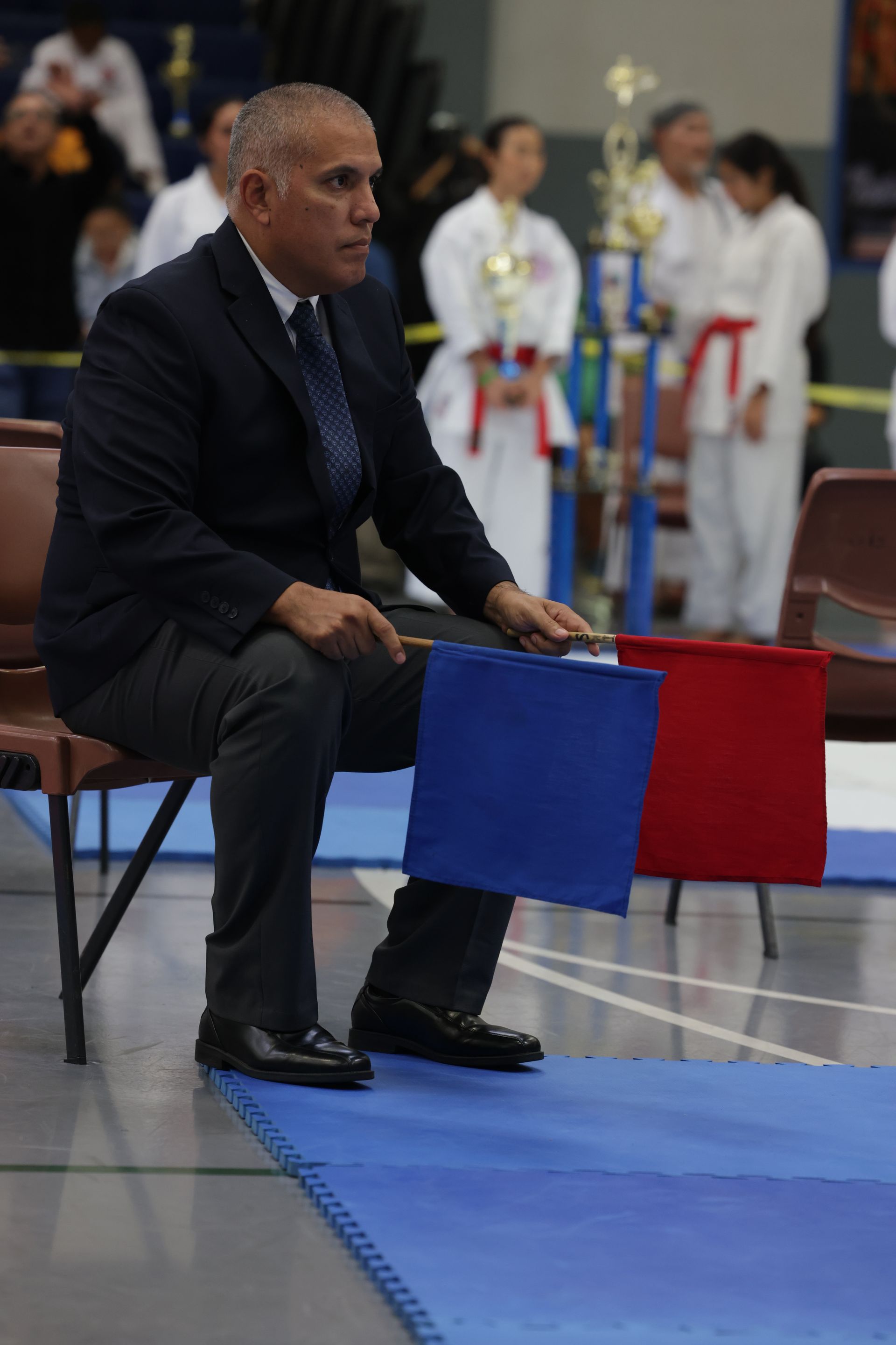 Official seated with red and blue flags at a martial arts event, competitors blurred in background