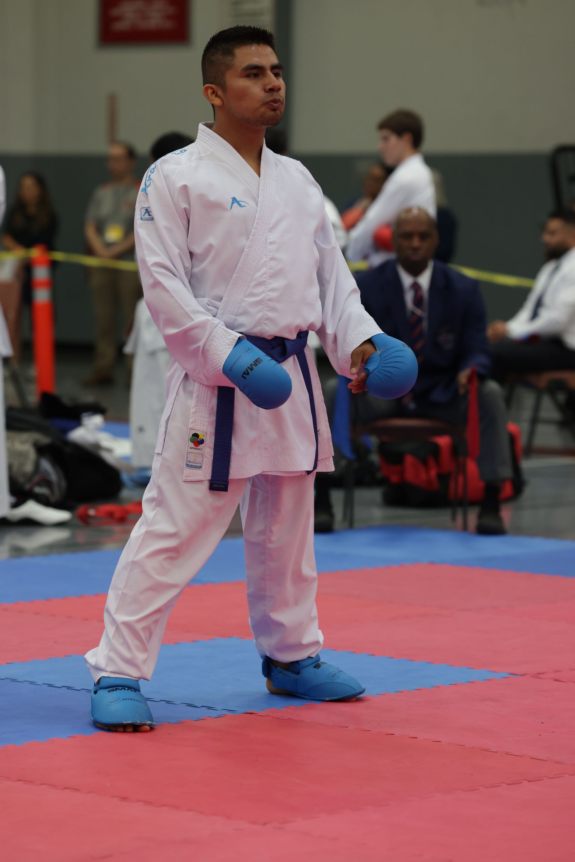 Karate competitor in white gi and blue gloves stands on a red mat in an indoor competition arena
