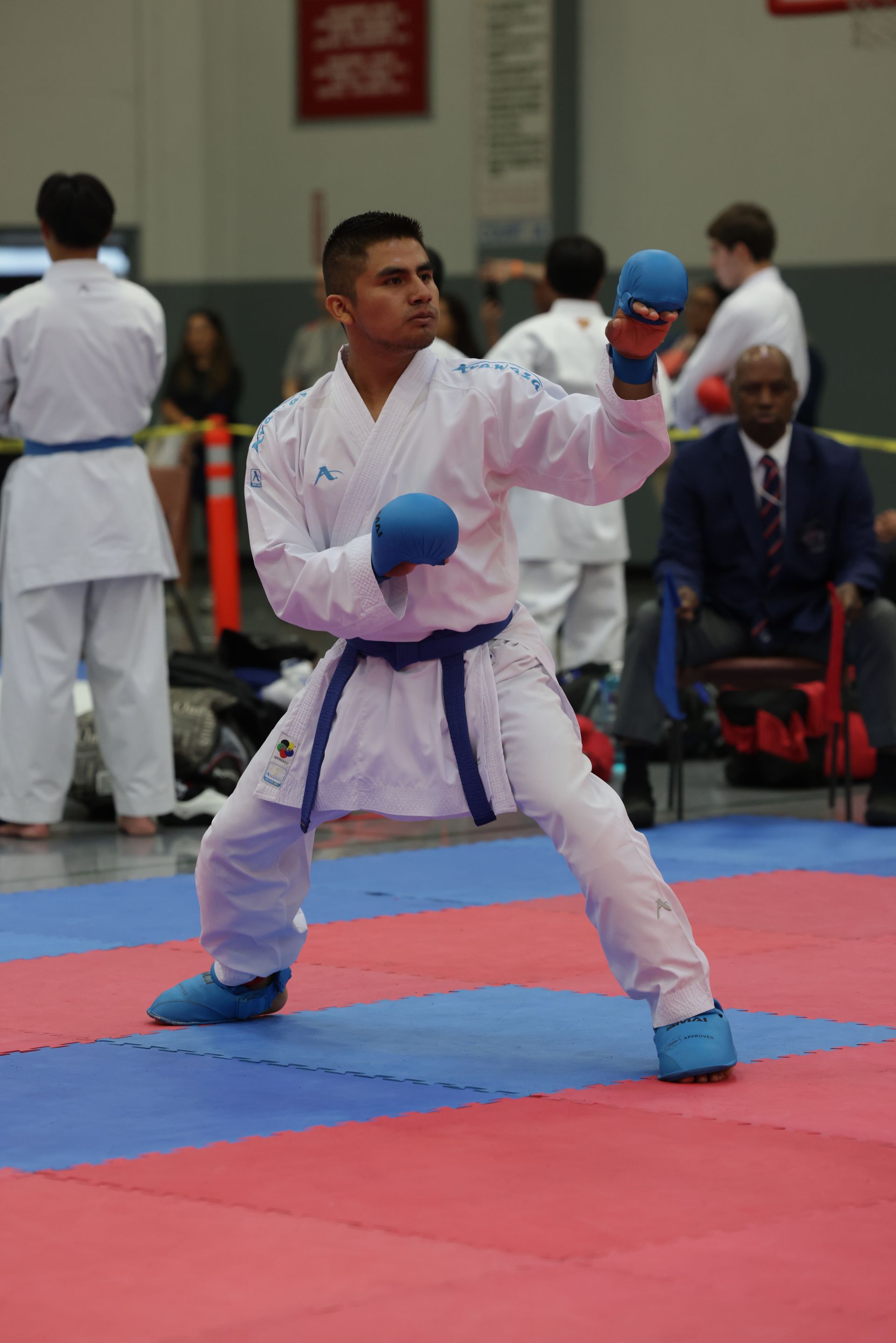 Karate competitor in a white gi and blue gloves performs a punch on a mat during a tournament