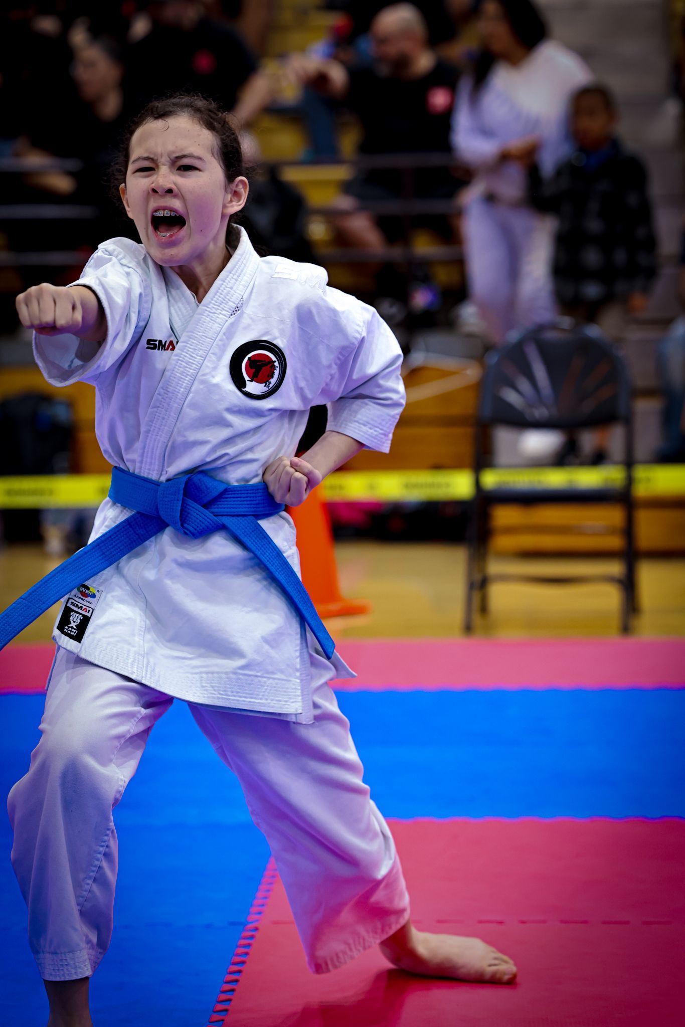 A karate practitioner in a white uniform and blue belt performs a punch with a focused expression on a competition mat.