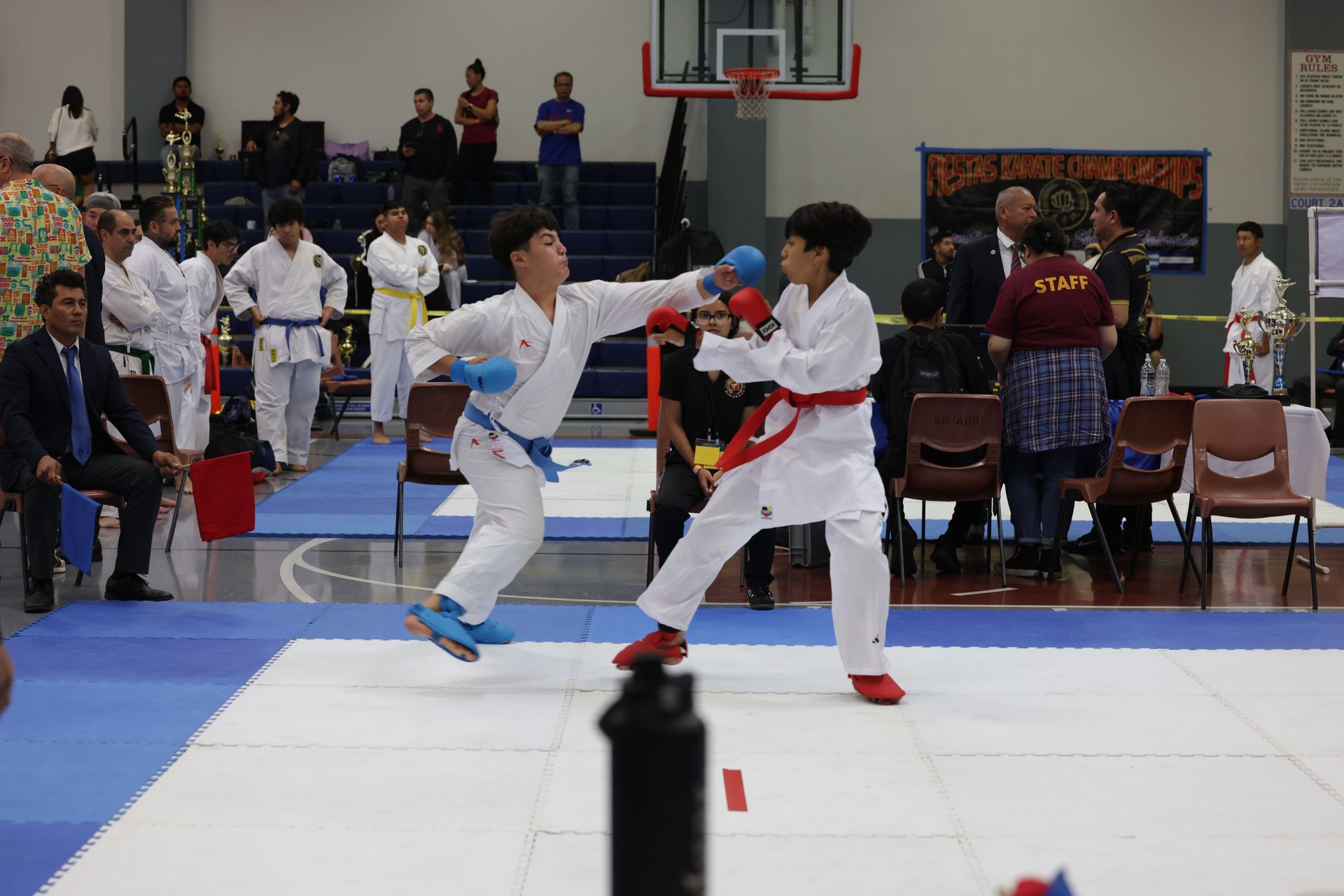 Two martial artists sparring on a competition mat in a gym, with coaches and spectators nearby.