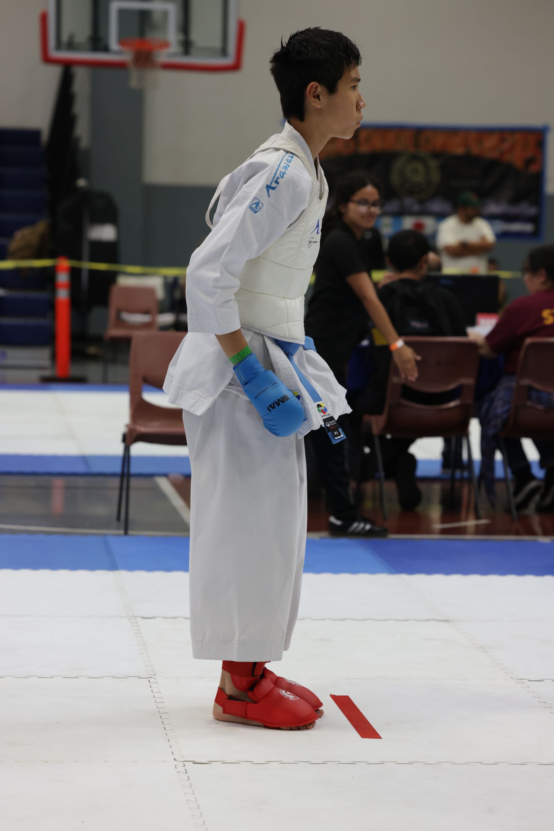 Karate competitor in white gi with blue gloves and red shoes stands on a mat in a gymnasium.