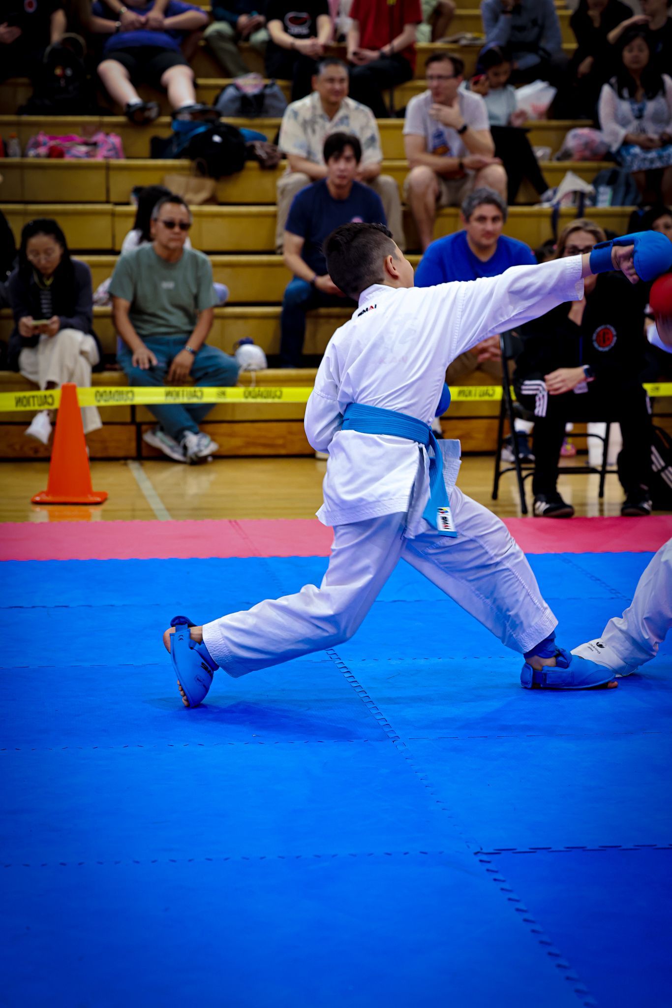 A karate competitor in a white uniform and blue belt performs a punch during a match on a blue mat in a gym.