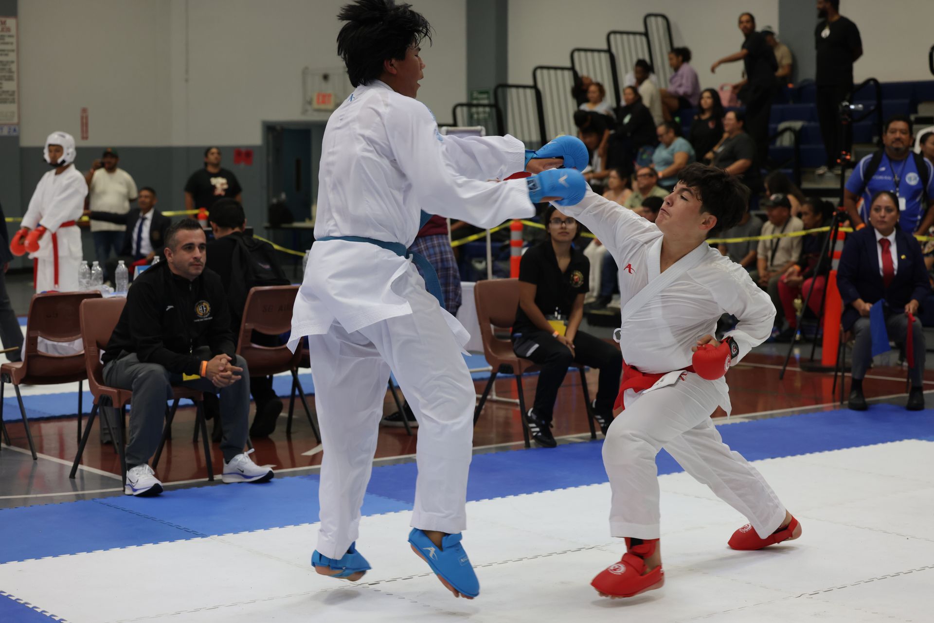 Two karate competitors sparring in a gym, one in white with blue gloves and one in white with red gloves