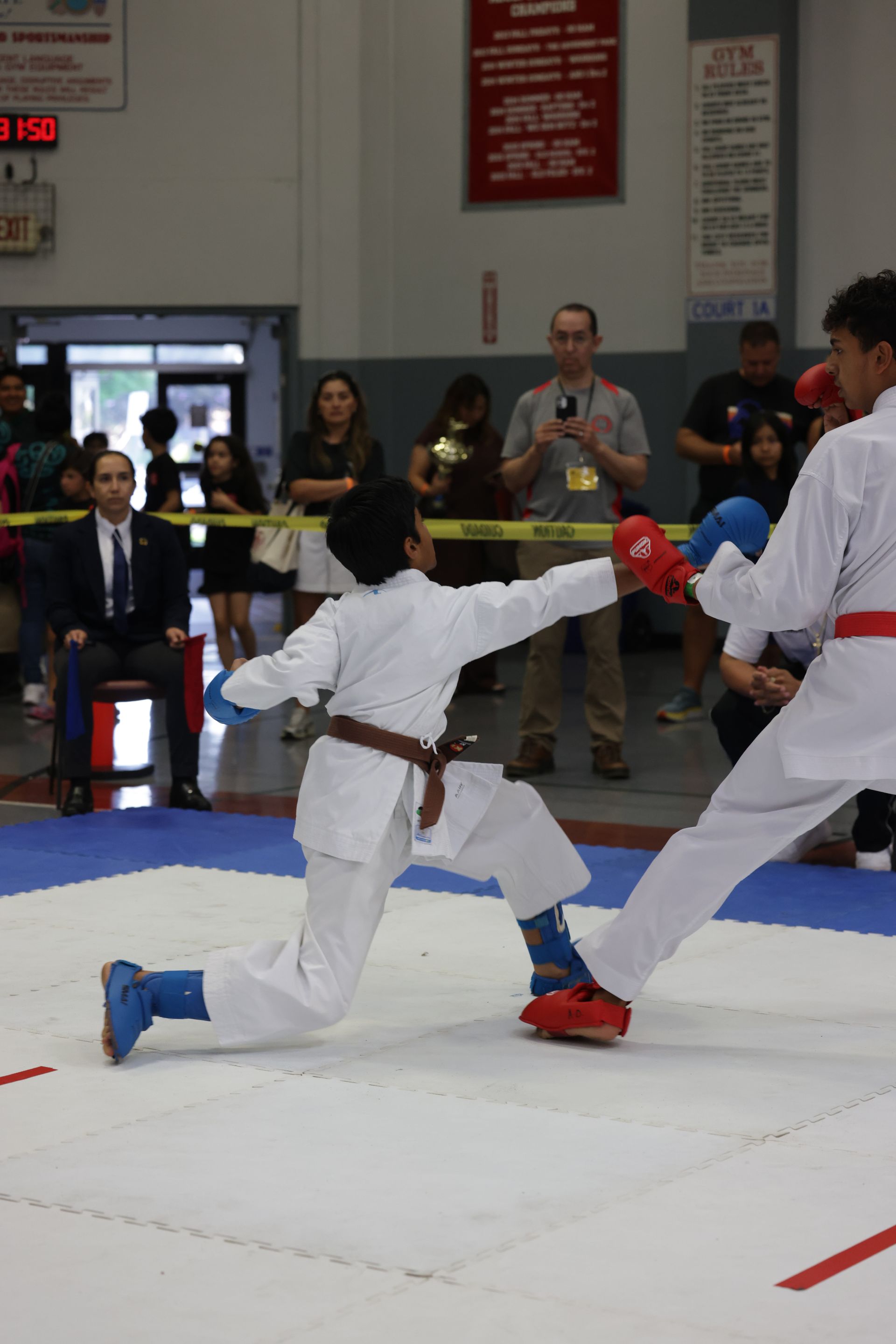 Two martial artists sparring on a mat, one kneeling and punching, in an indoor competition setting