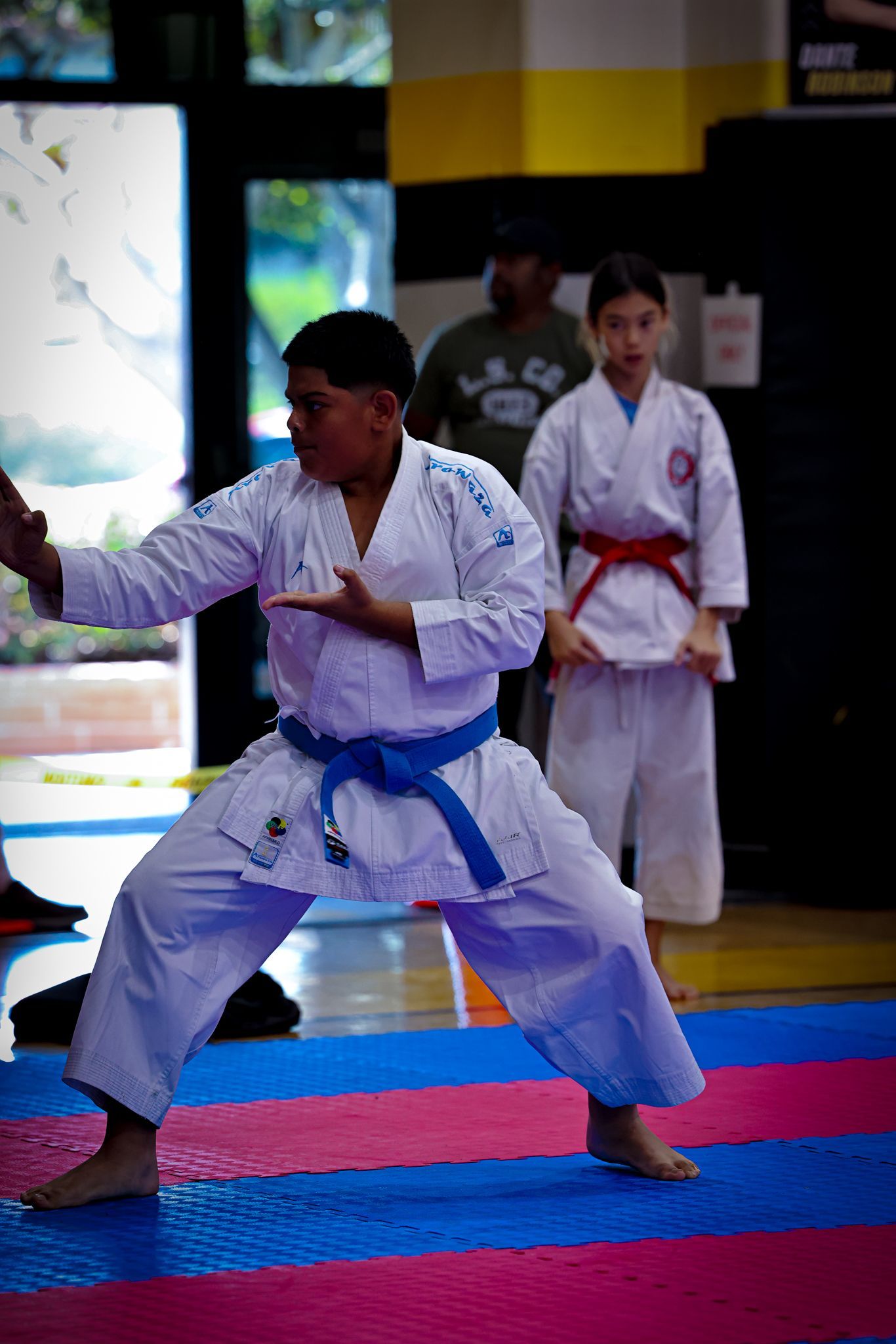 Two martial arts students in white uniforms practice a stance on a blue and pink matted gym floor.