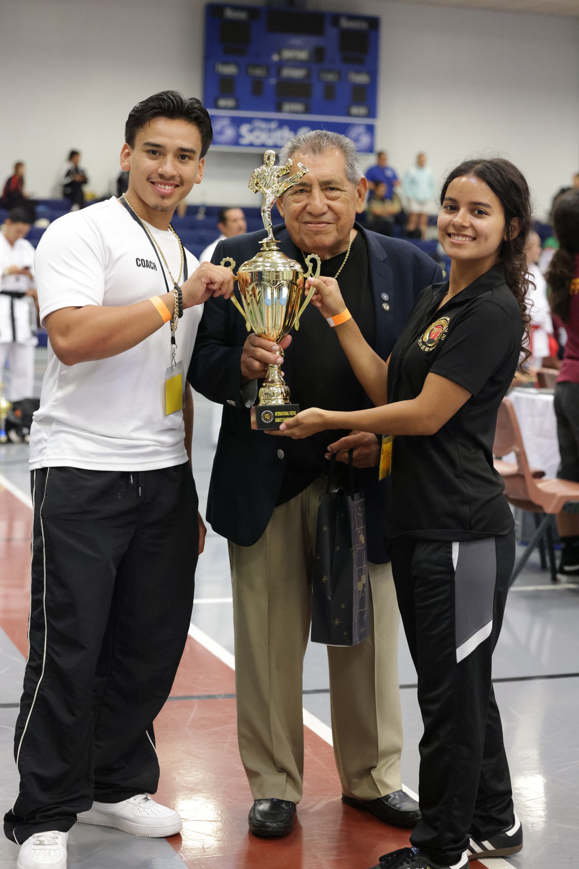 Three people hold a trophy together in a gymnasium during a sports event.