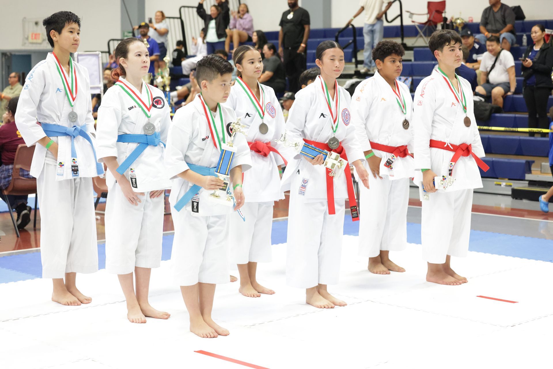 Karate competitors in white uniforms and colored belts standing in line at an indoor gymnasium