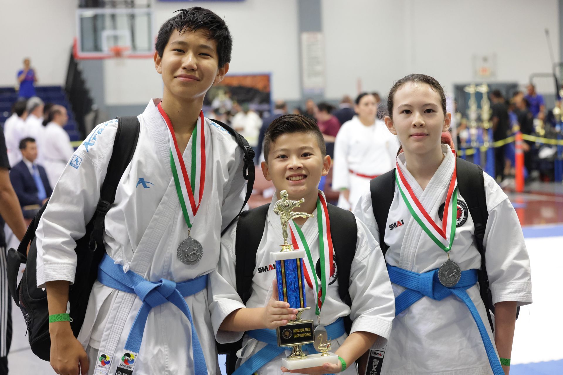 Three martial arts competitors posing with medals and a trophy at an indoor event