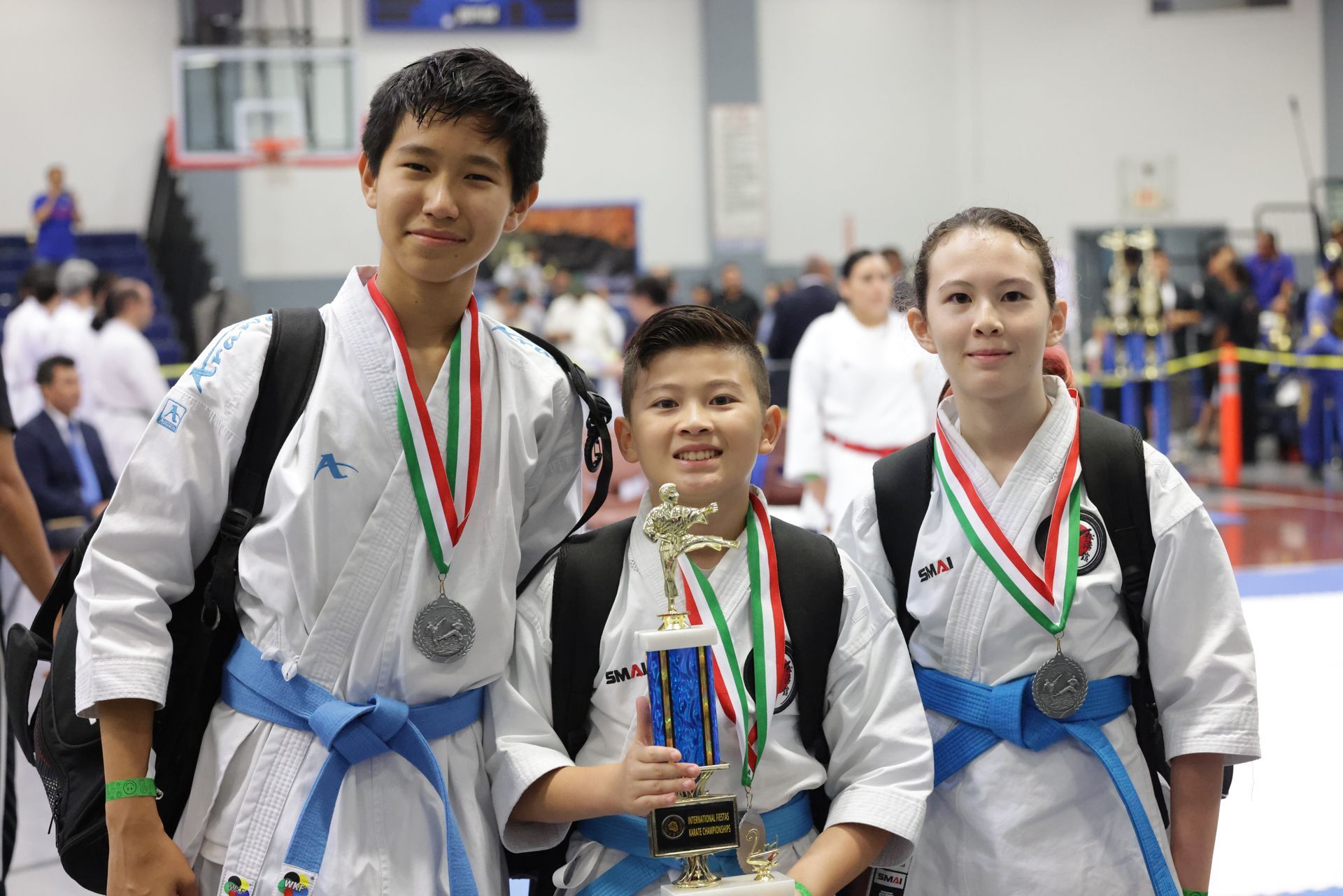 Three martial arts medalists in white uniforms posing indoors, with blue, silver, and red medals.