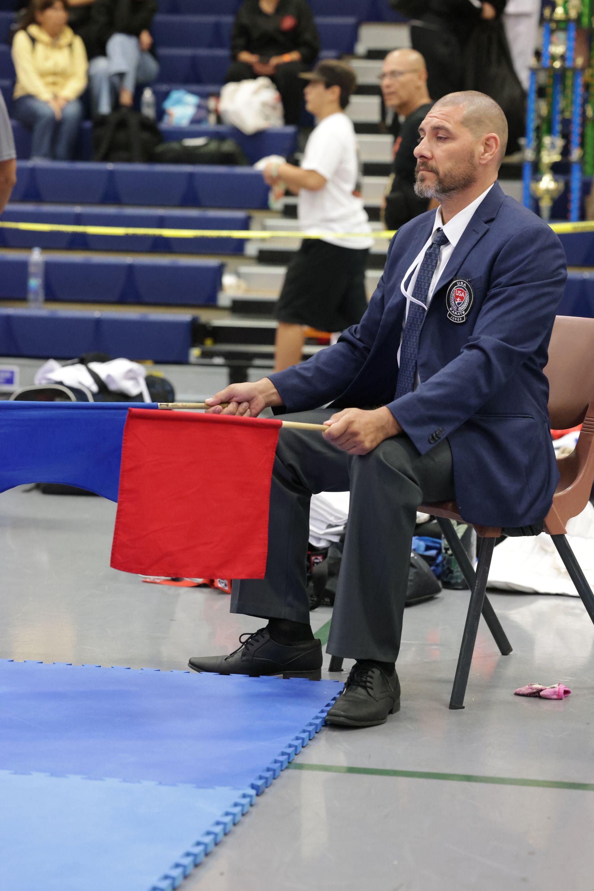 Man seated courtside in a navy blazer holding a red flag at a sports event