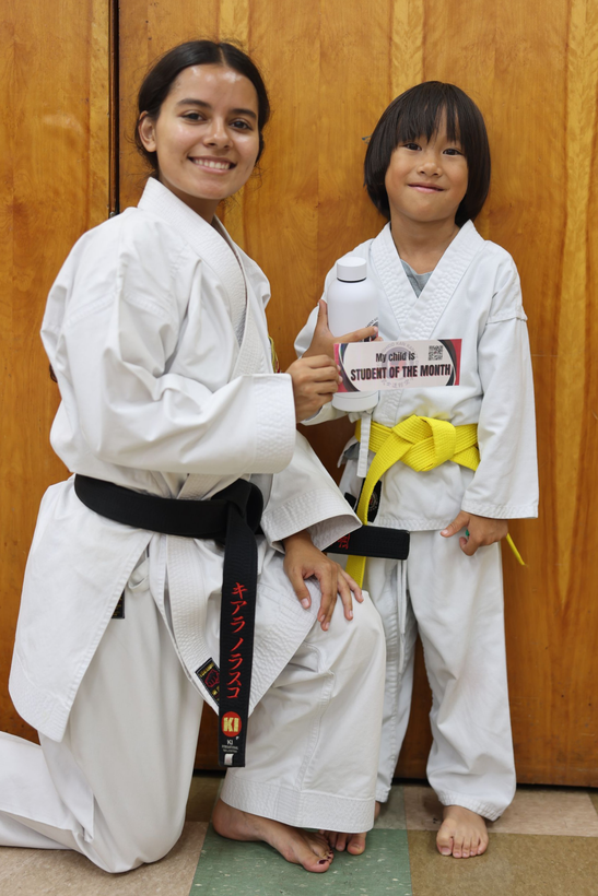 Two martial arts practitioners in white gis pose with belts in front of a wooden door.