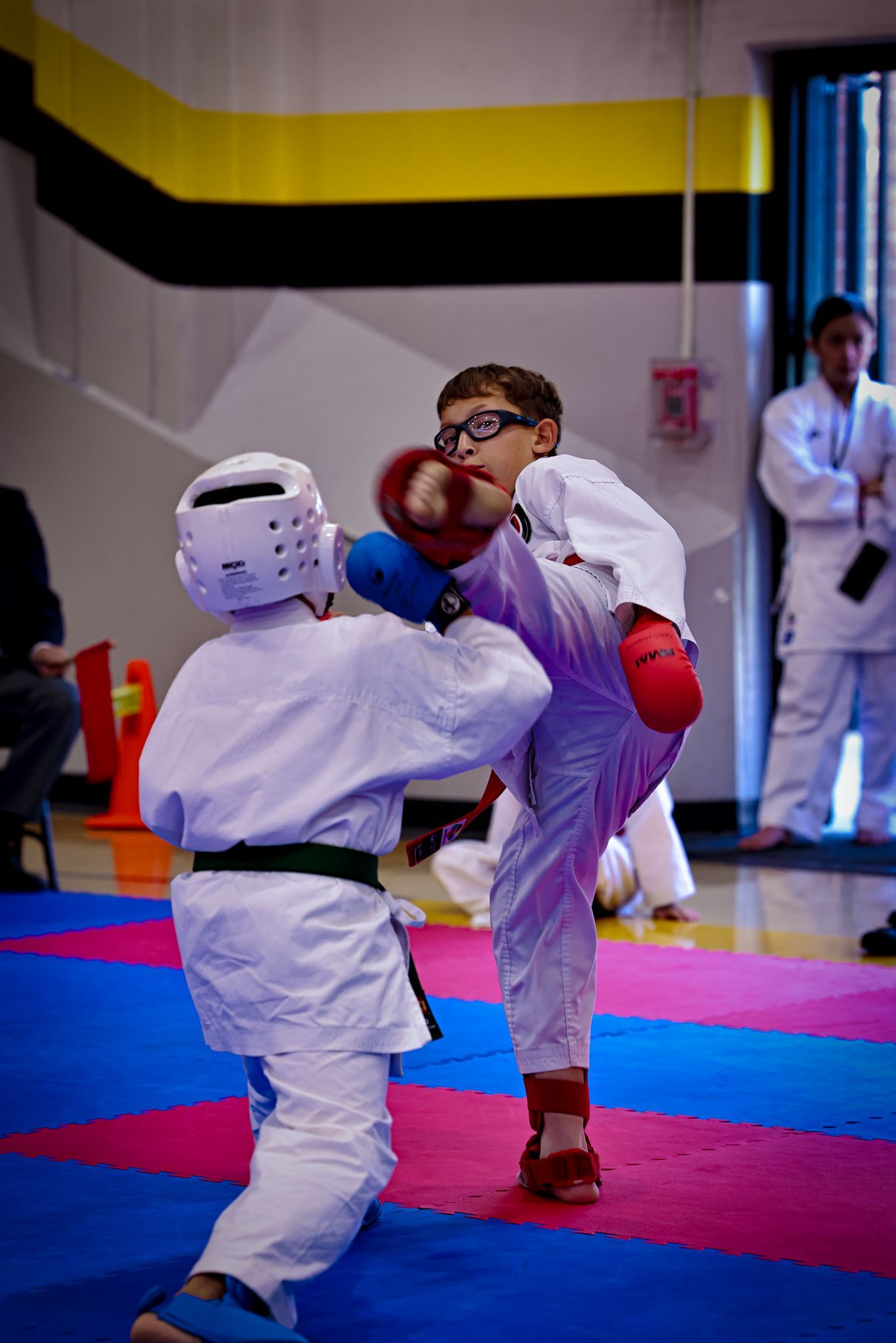 Two martial artists in white uniforms sparring on a blue and pink mat; one performs a high kick while wearing red gear.