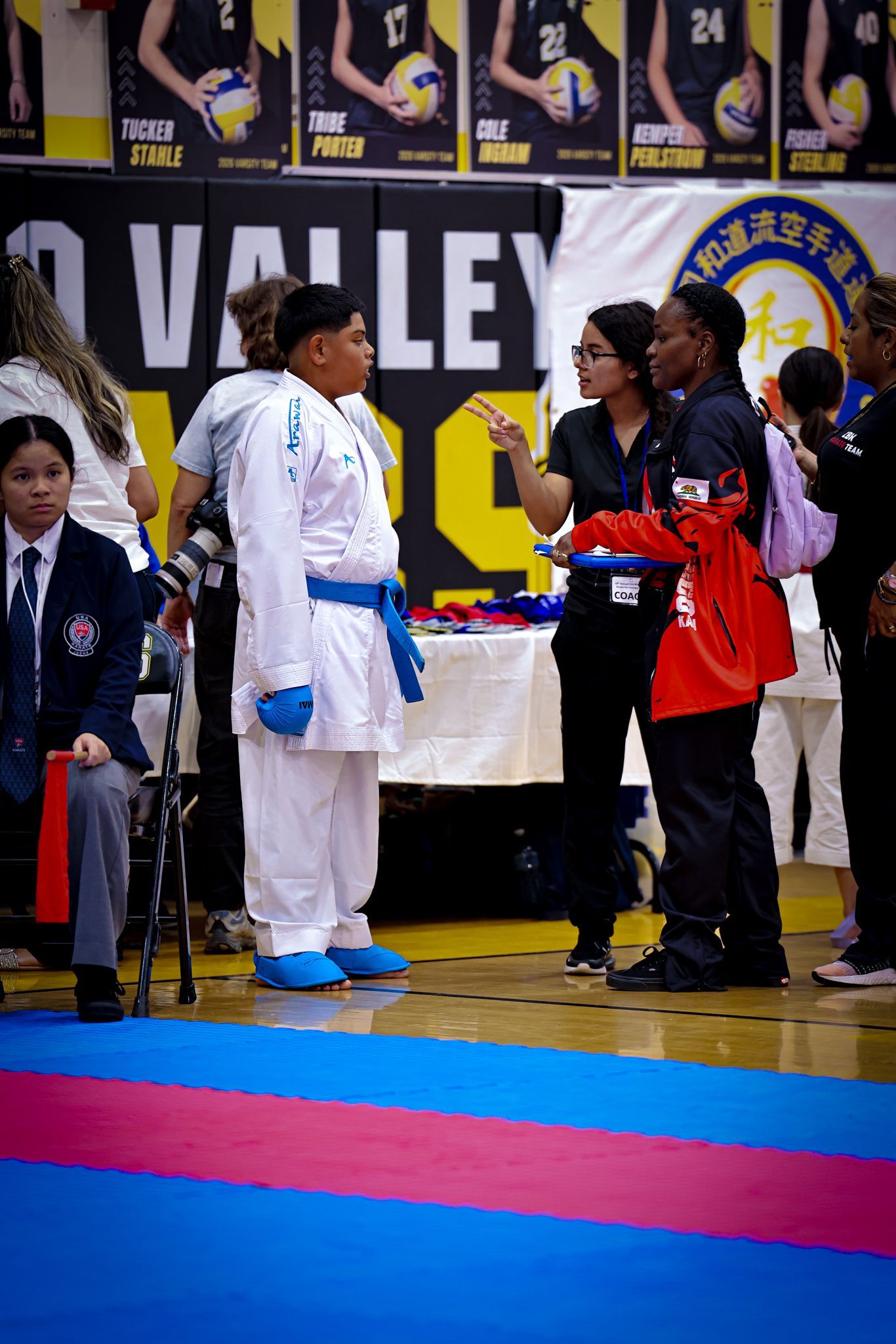 A person in a white karate uniform with a blue belt stands and talks to two people at a sports tournament.