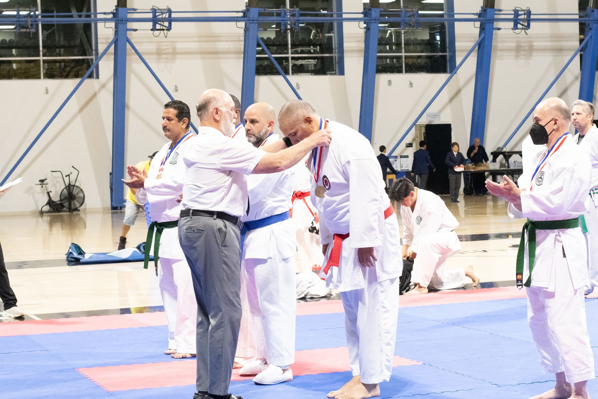 Man receiving a medal at a karate tournament, indoors. Other karate participants watch.
