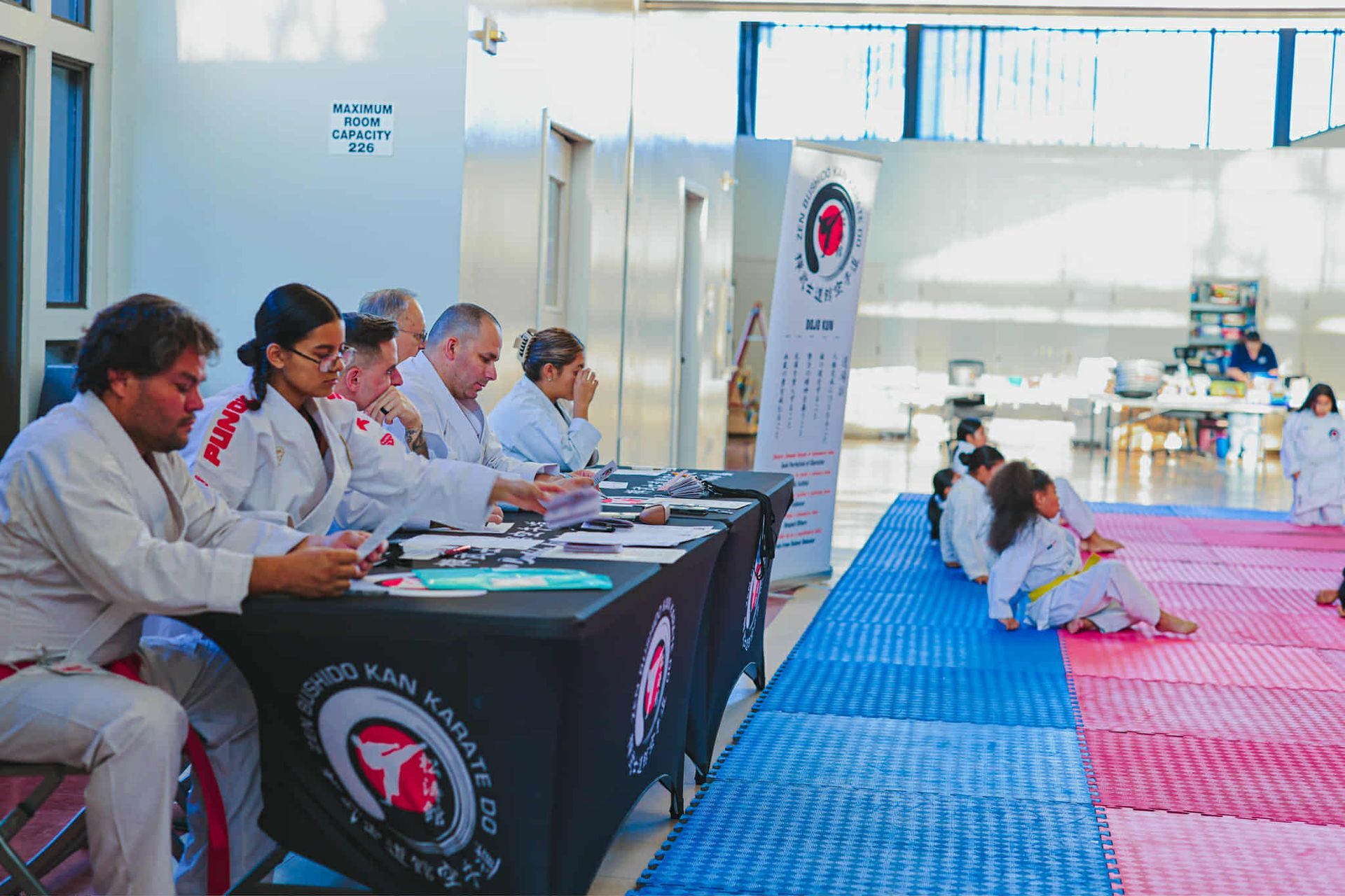 Karate judges at a competition; seated, reviewing notes. Competitors in the background on mats.