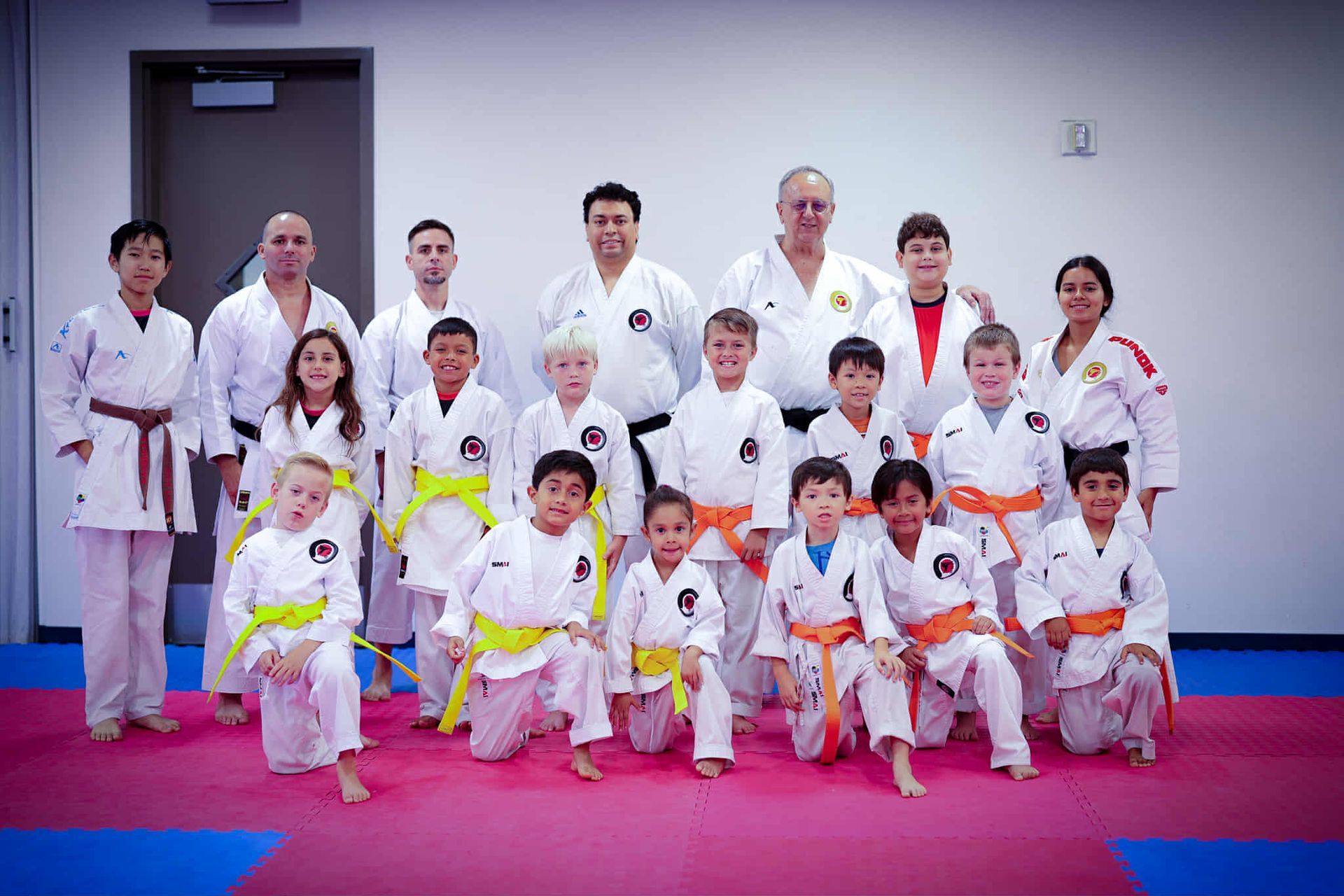 Karate class photo: group of children and adults in white uniforms, colored belts, posing on a red and blue mat.
