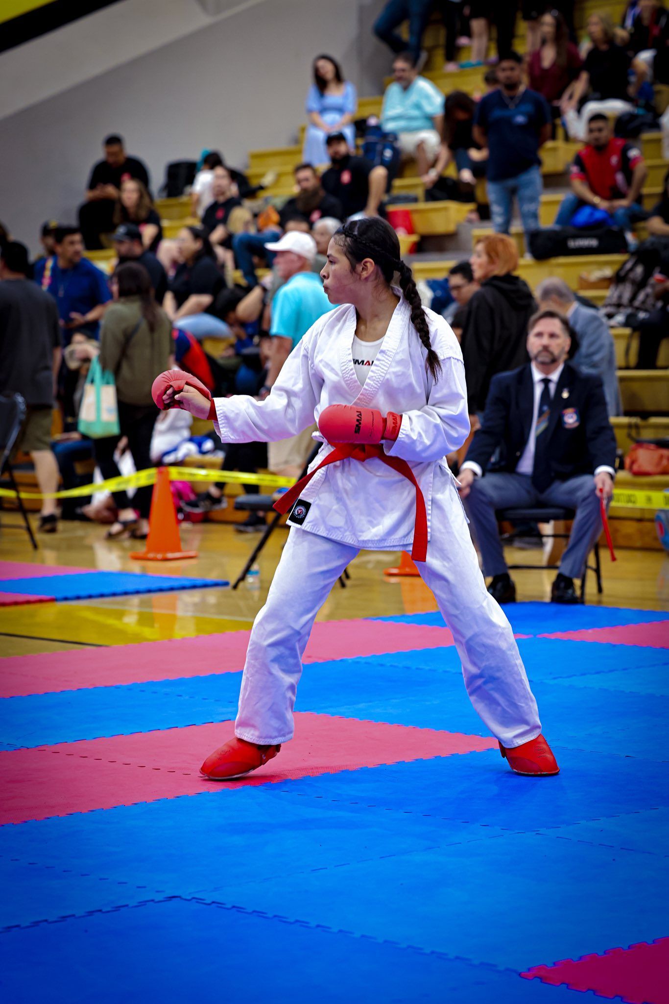 A martial artist in a white gi with red gear stands in a defensive stance on a gym mat during a tournament.