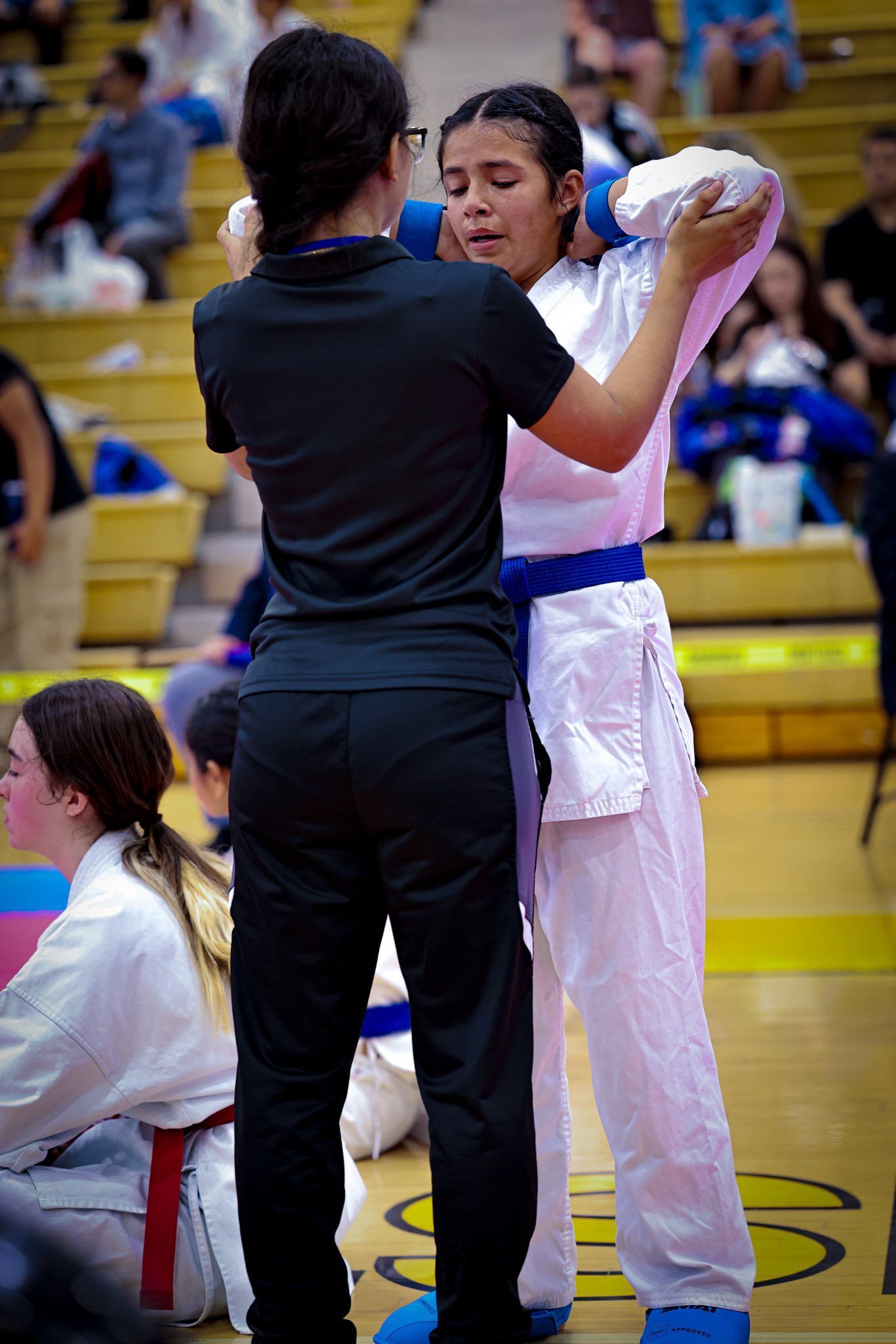 A person in a black uniform assists a karate practitioner in a white gi and blue belt with their headgear in a gym.