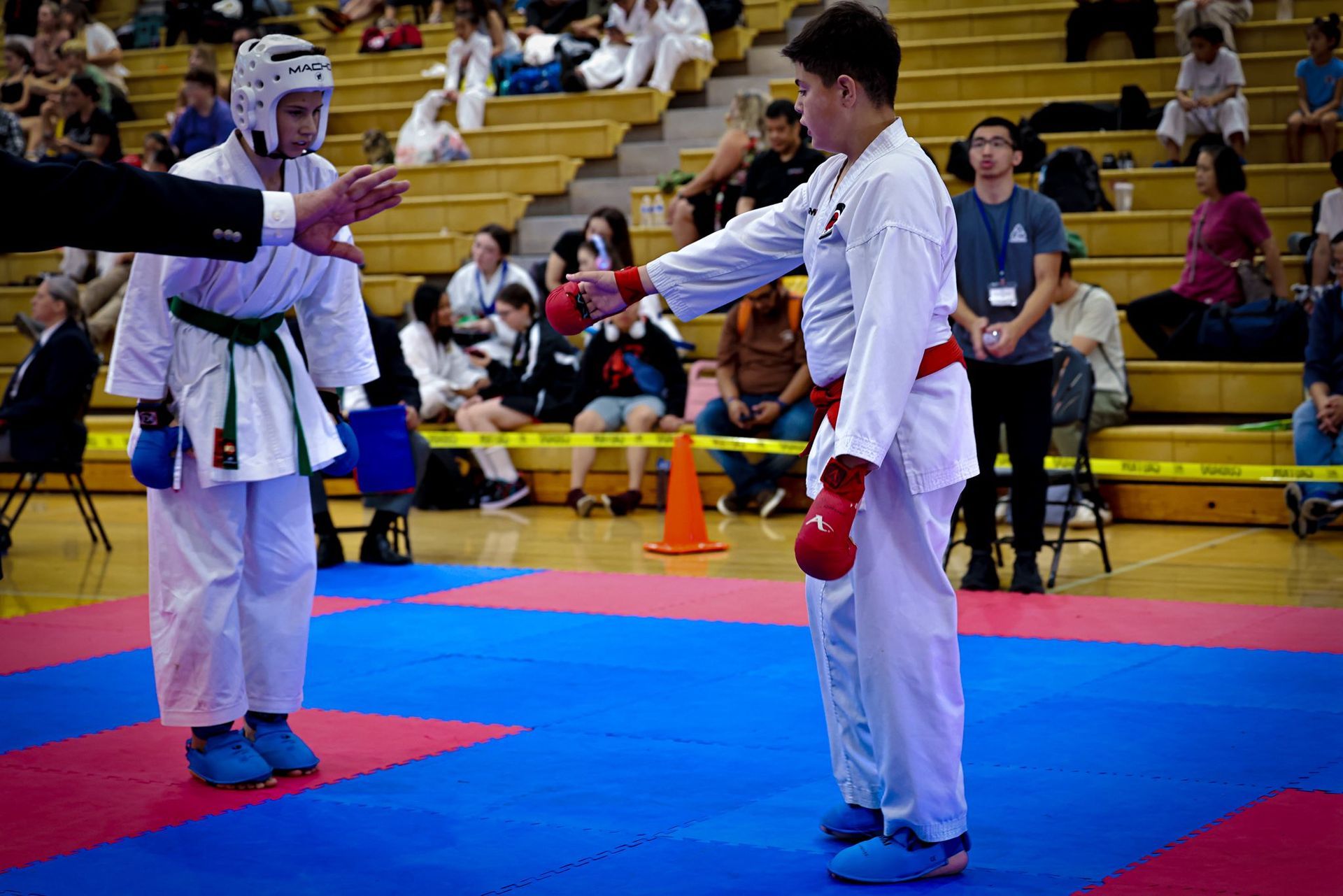 Two karate practitioners in white uniforms bow to each other on a blue and pink mat in a gymnasium.