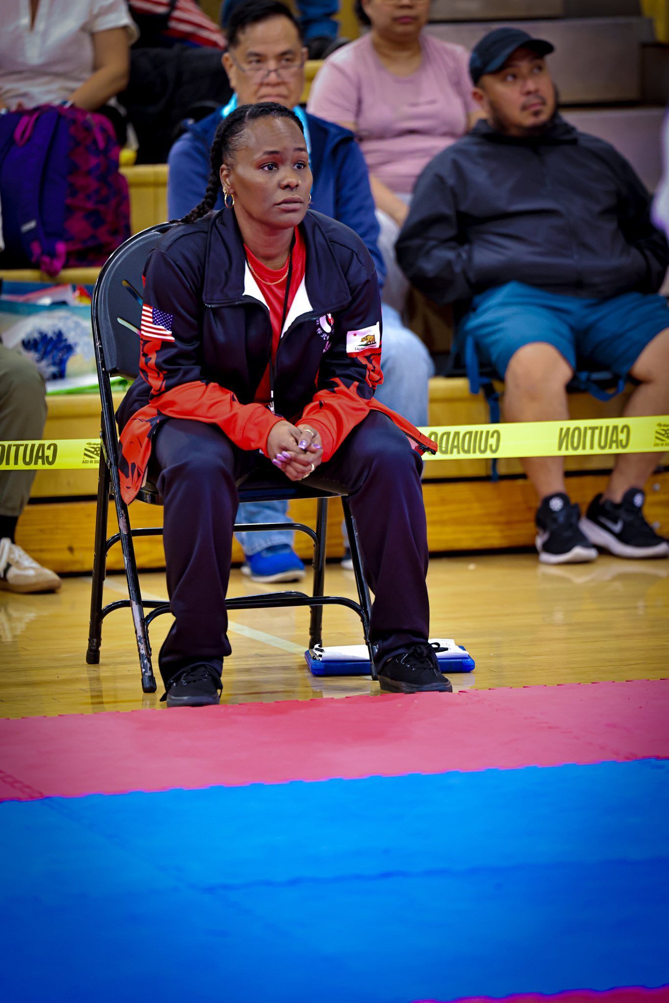 A person in a black and red warm-up suit sits on a folding chair in an indoor gym, watching an athletic competition.