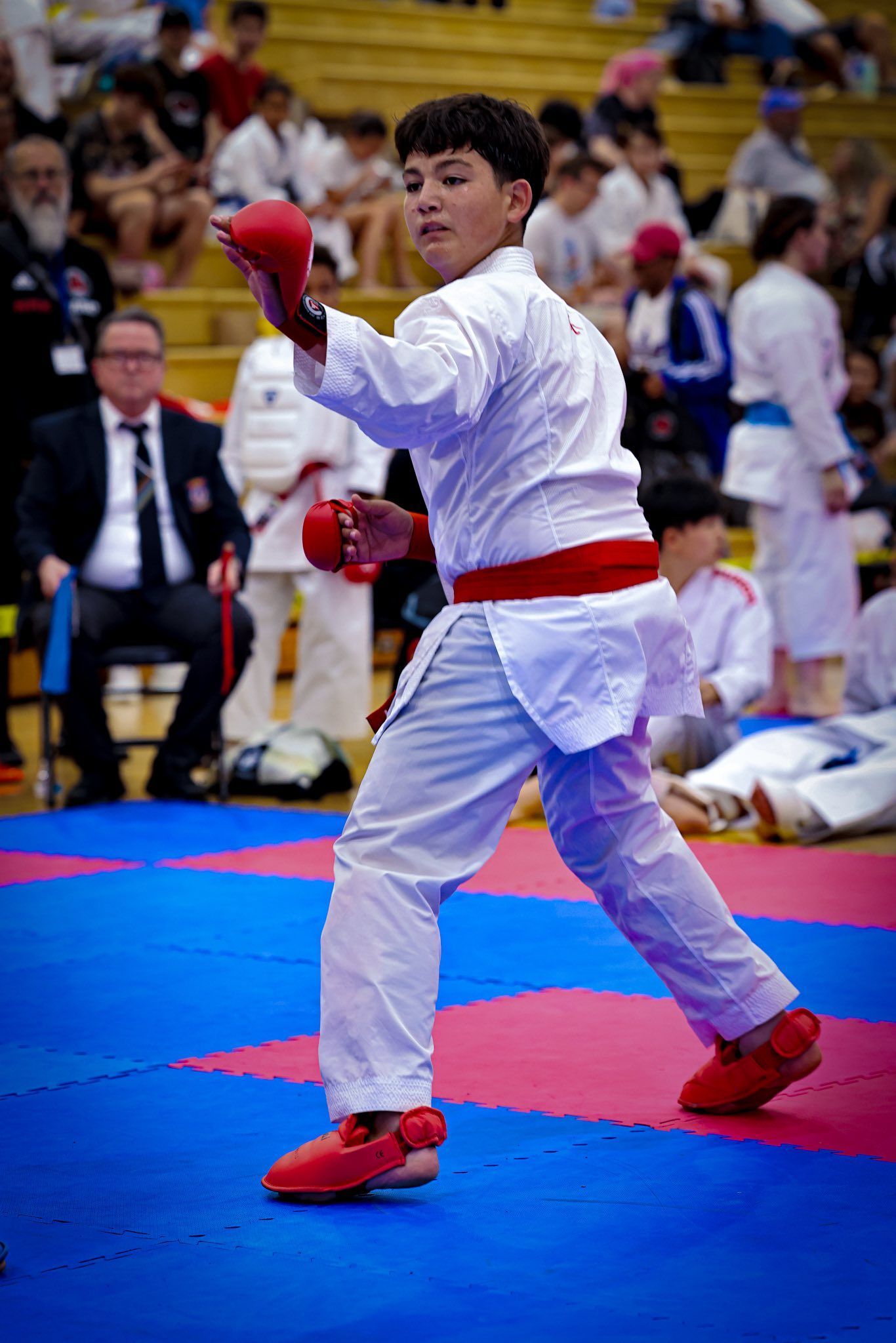 A karate competitor in a white uniform with red gear performs a move on a red and blue mat in a gym with spectators.