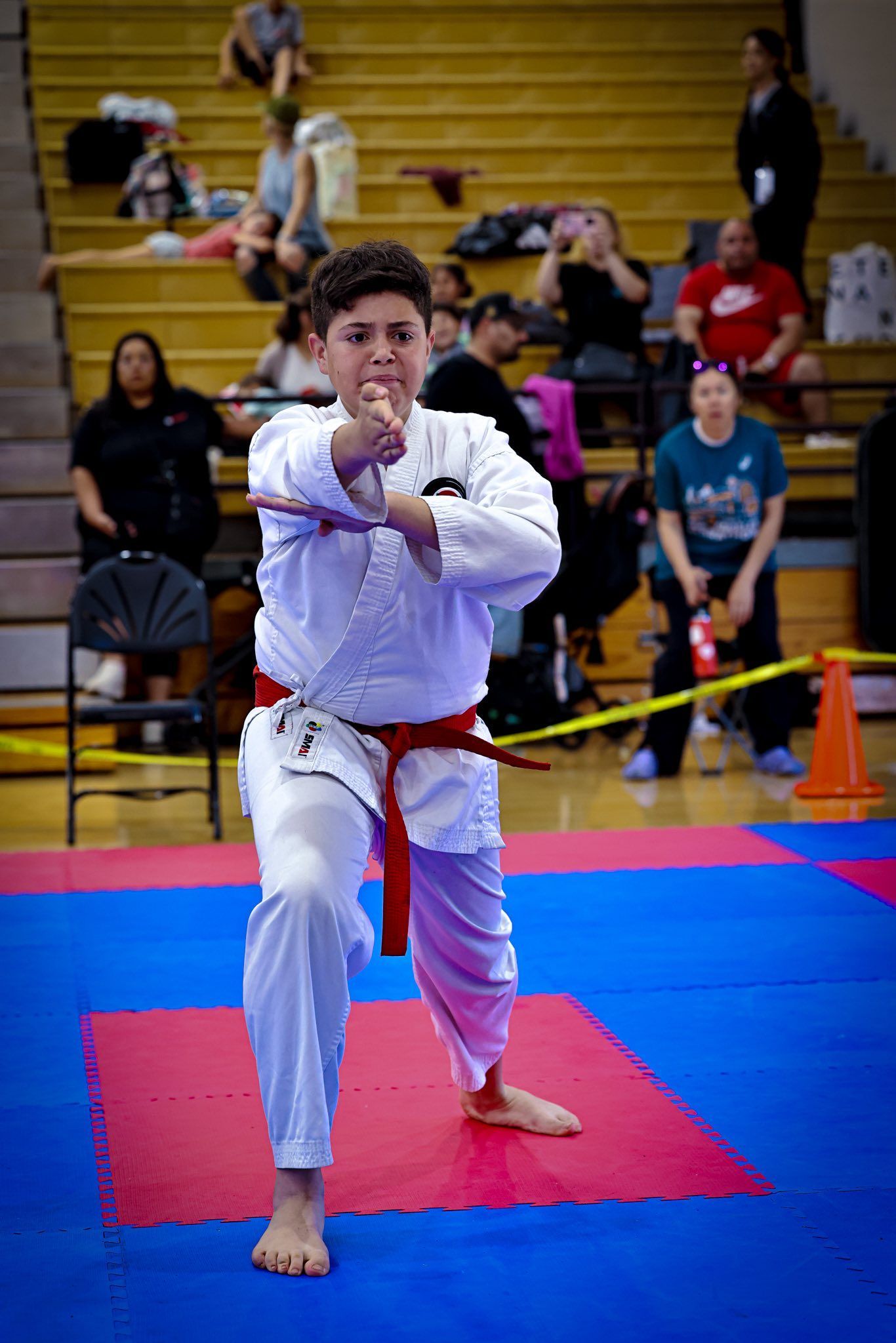 A martial arts practitioner in a white gi and red belt performs a kata pose on a blue and red mat in a gymnasium.