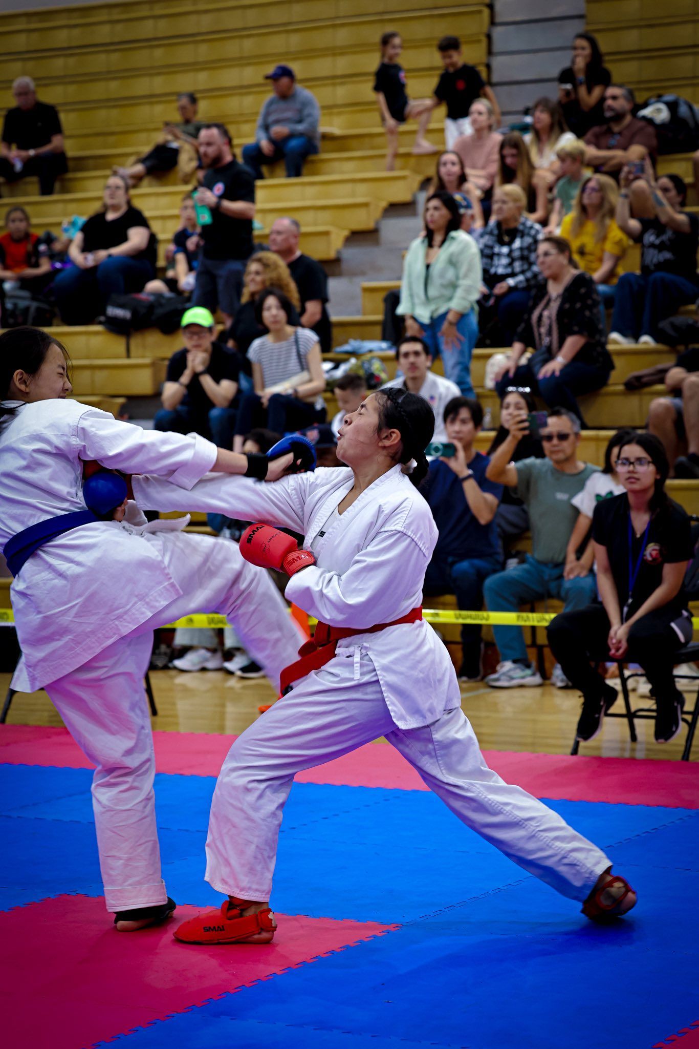 Two martial artists in white uniforms sparring on a blue and pink mat in front of a seated audience.
