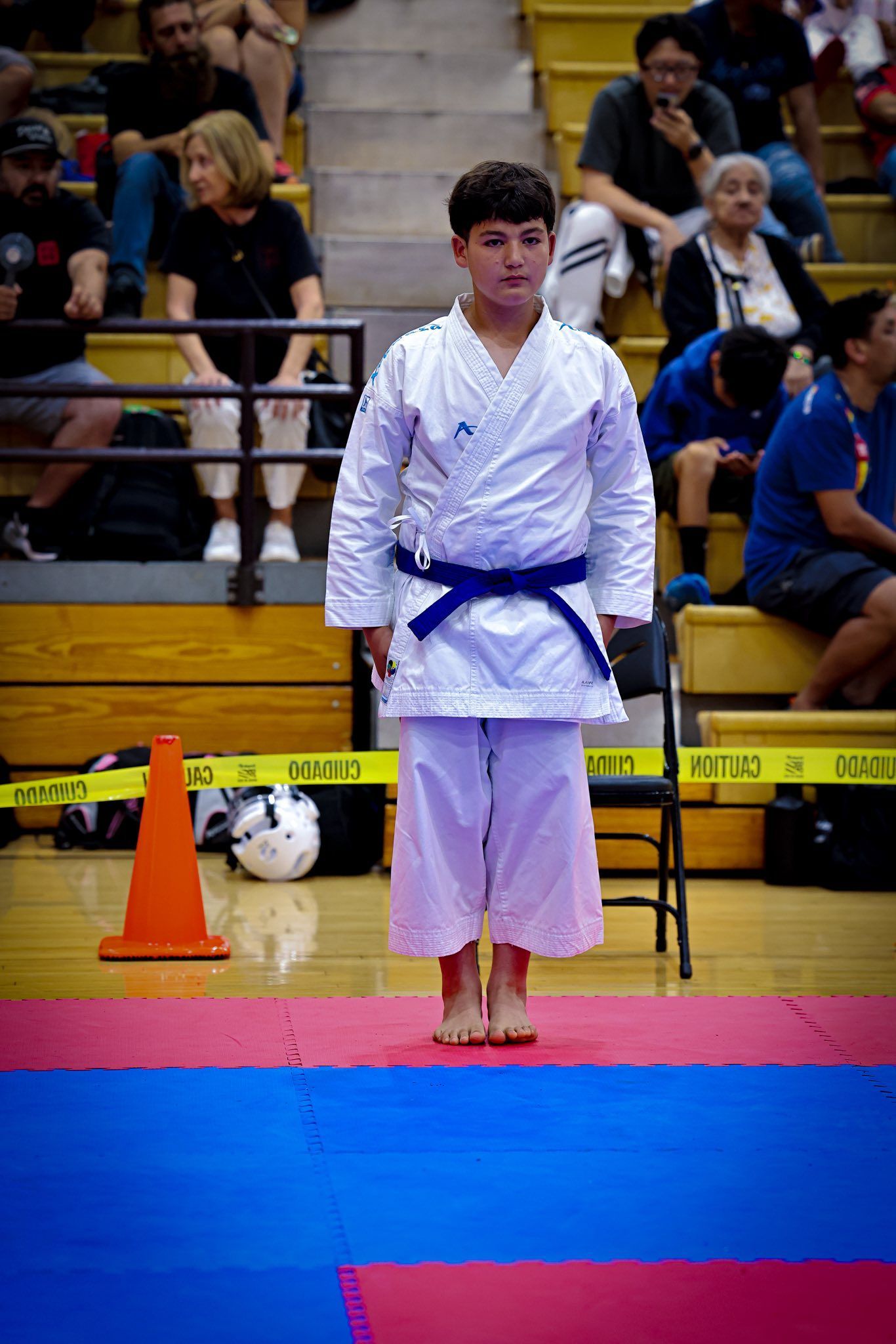 A person in a white karate uniform with a blue belt stands centered on a blue and pink mat in a gymnasium.