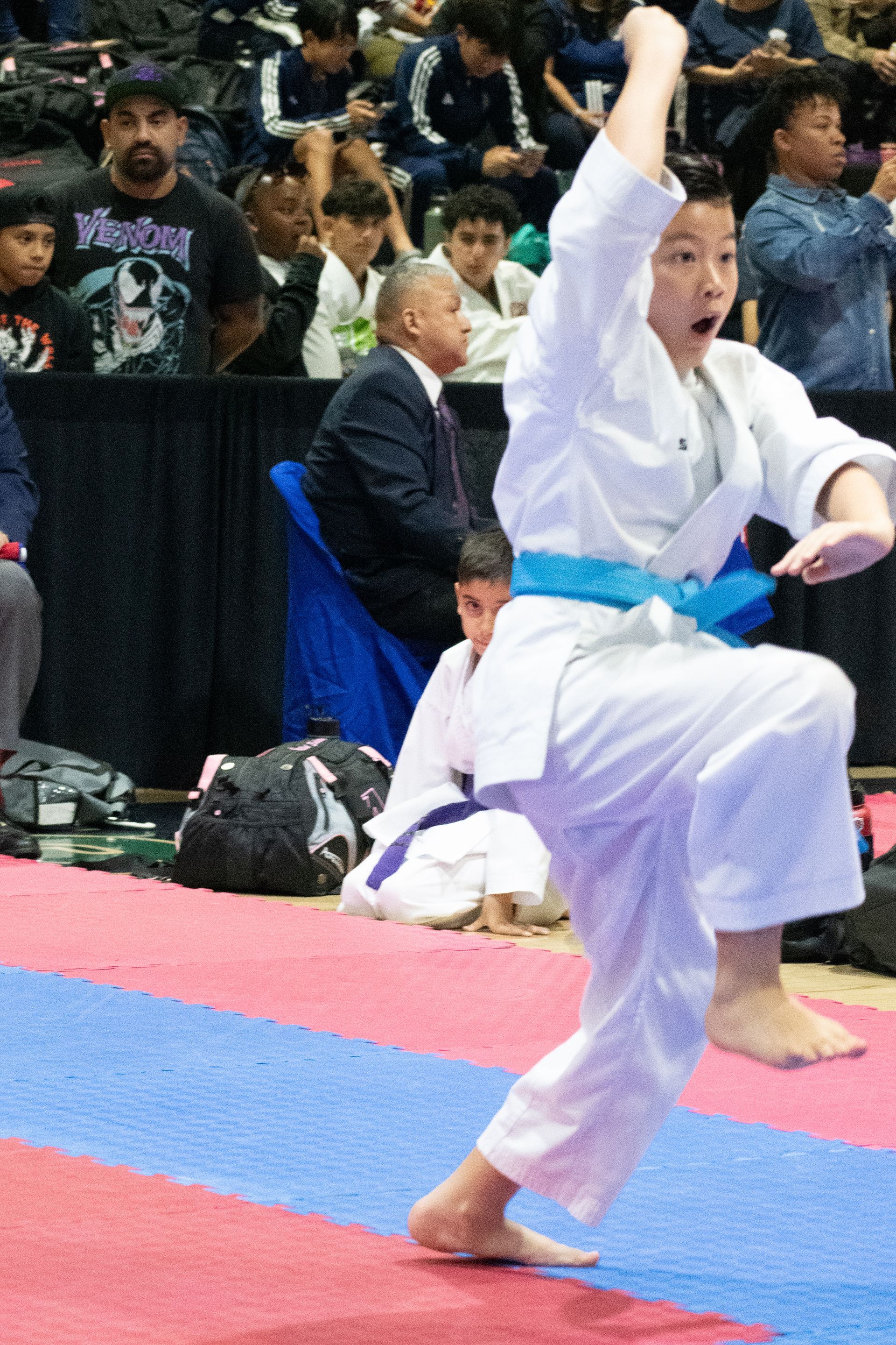 Boy in a white karate uniform performs a high kick with blue belt on a competition mat.