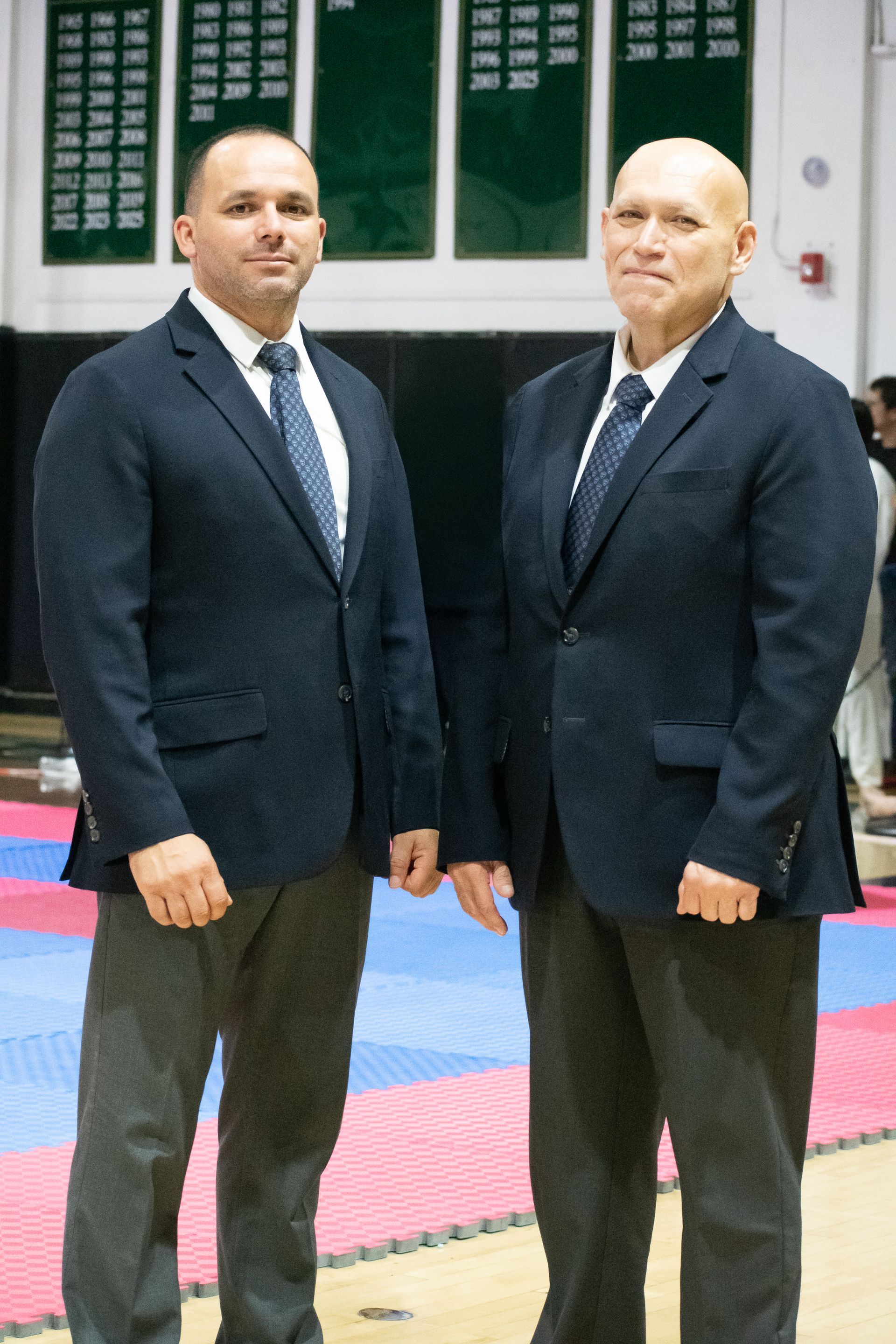 Two men in blazers and ties standing side by side in a gymnasium setting. They are in front of a competition mat.