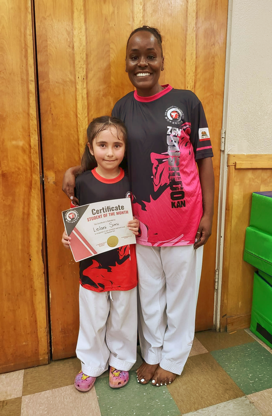 Two girls standing together in front of wooden doors, smiling and holding a certificate.