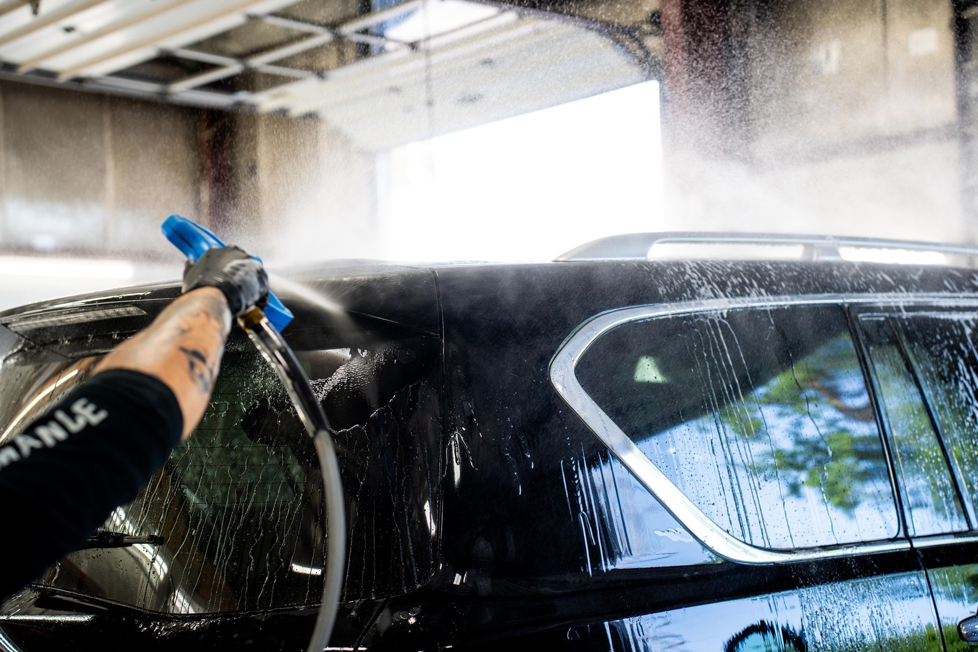 Person pressure-washing a black car in a garage, spraying water over the rear window