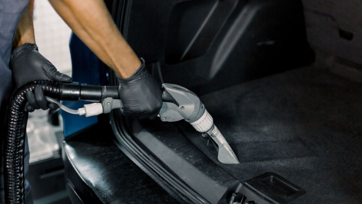 Gloved hand using a vacuum nozzle to clean a car’s black carpeted floor mat and door sill area
