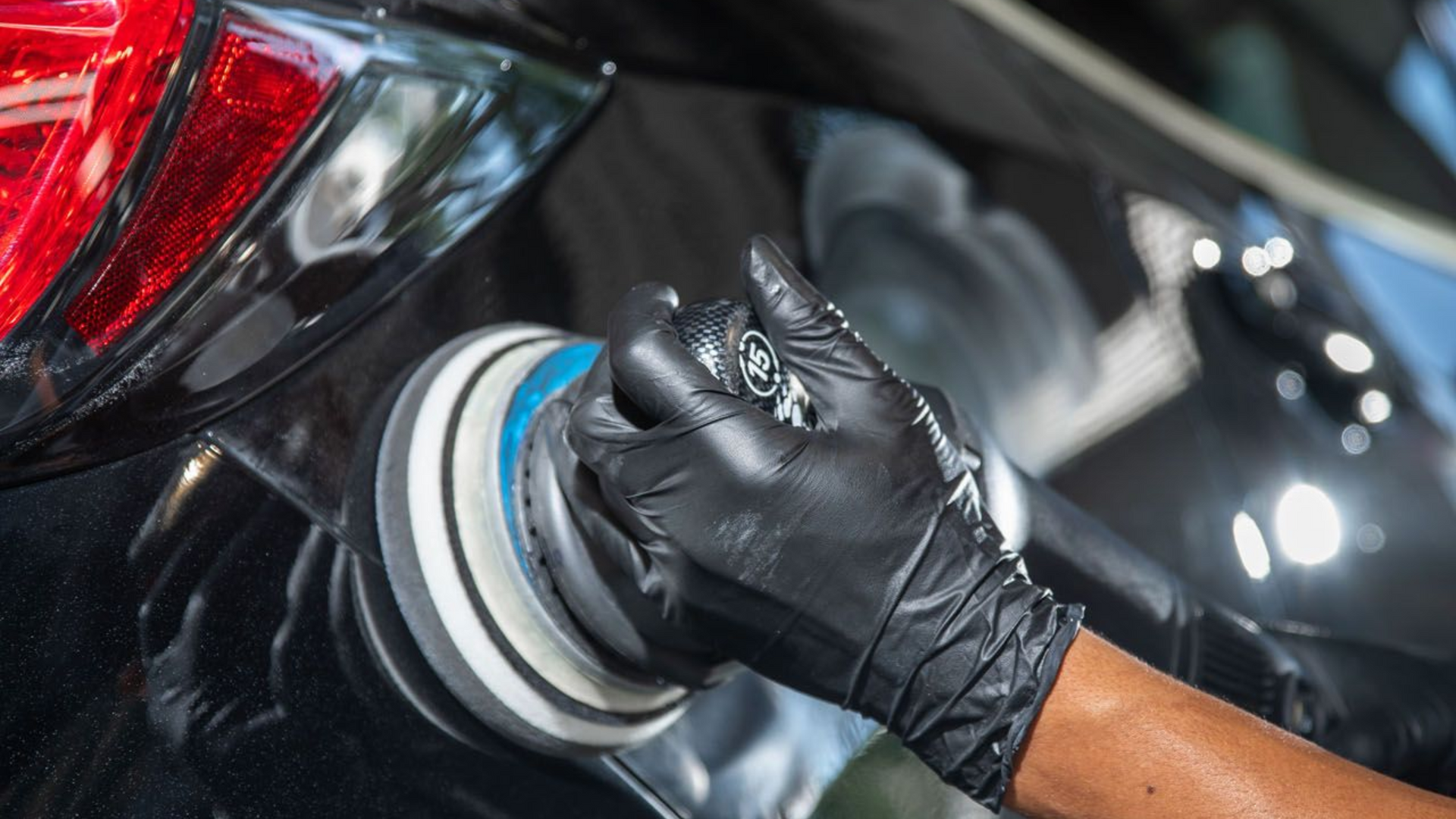 Gloved hand polishing a black car with a buffing pad, close-up on the rear quarter panel.