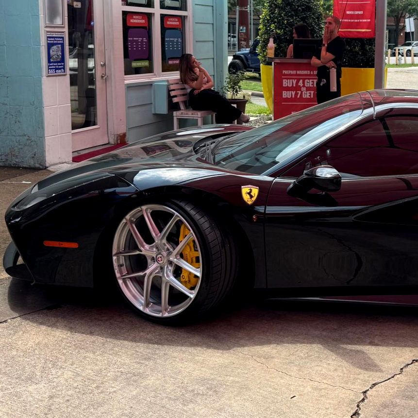 Black Ferrari parked outside a storefront, with a person sitting nearby on a bench.