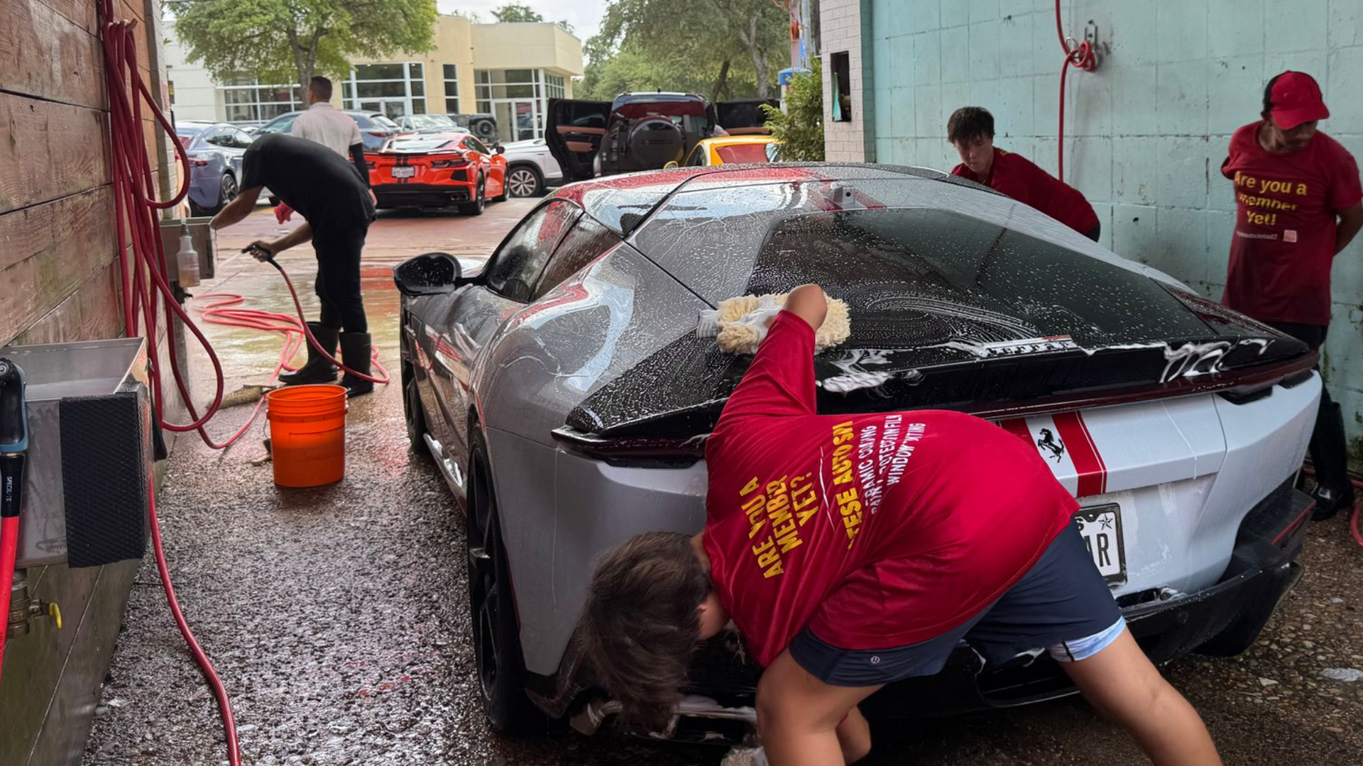 People hand-wash a silver car under a covered area, with buckets, suds, and wet pavement nearby.